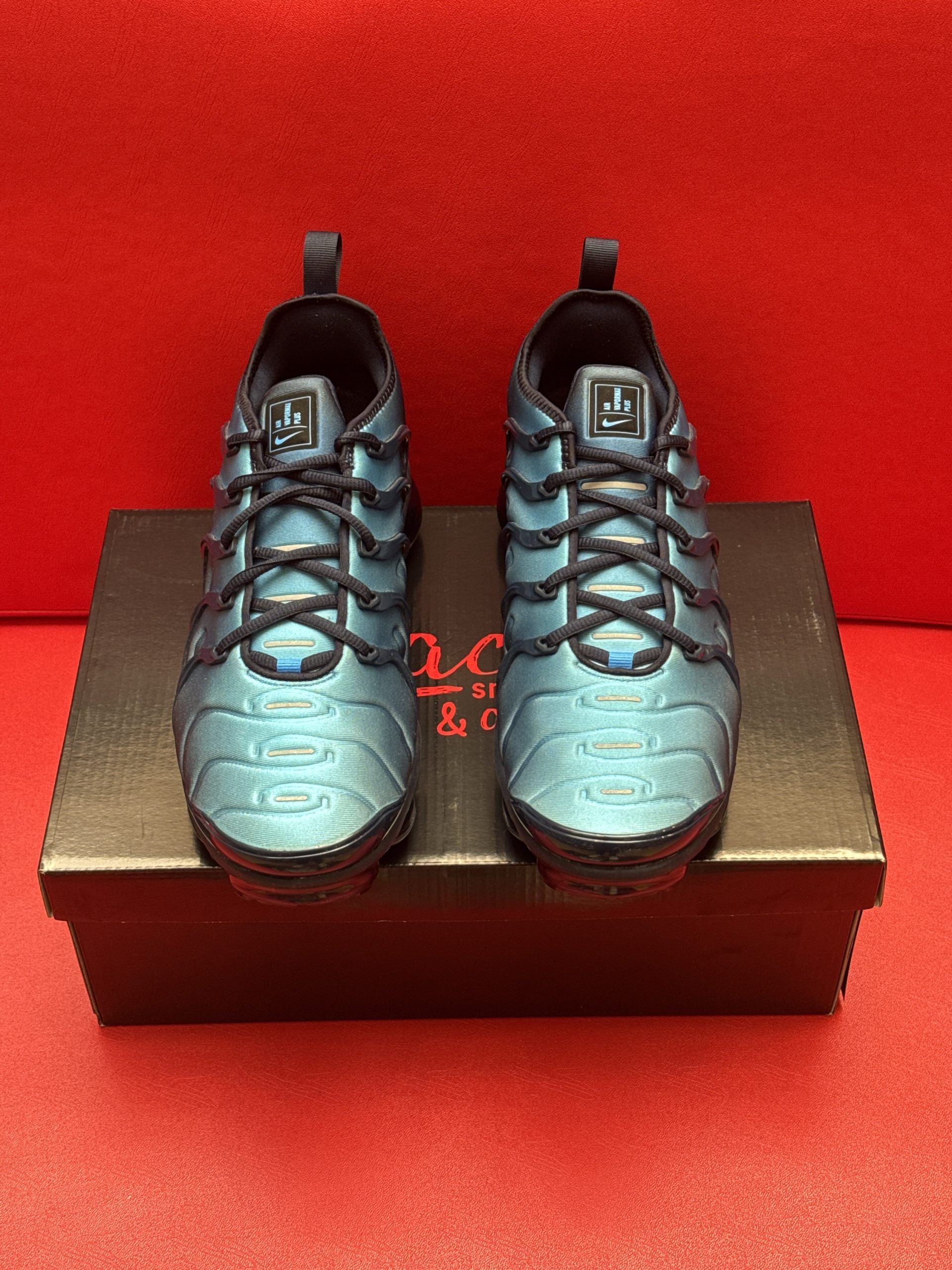 Blue and black Nike Air VaporMax Plus sneakers on a black shoebox, against a red backdrop.