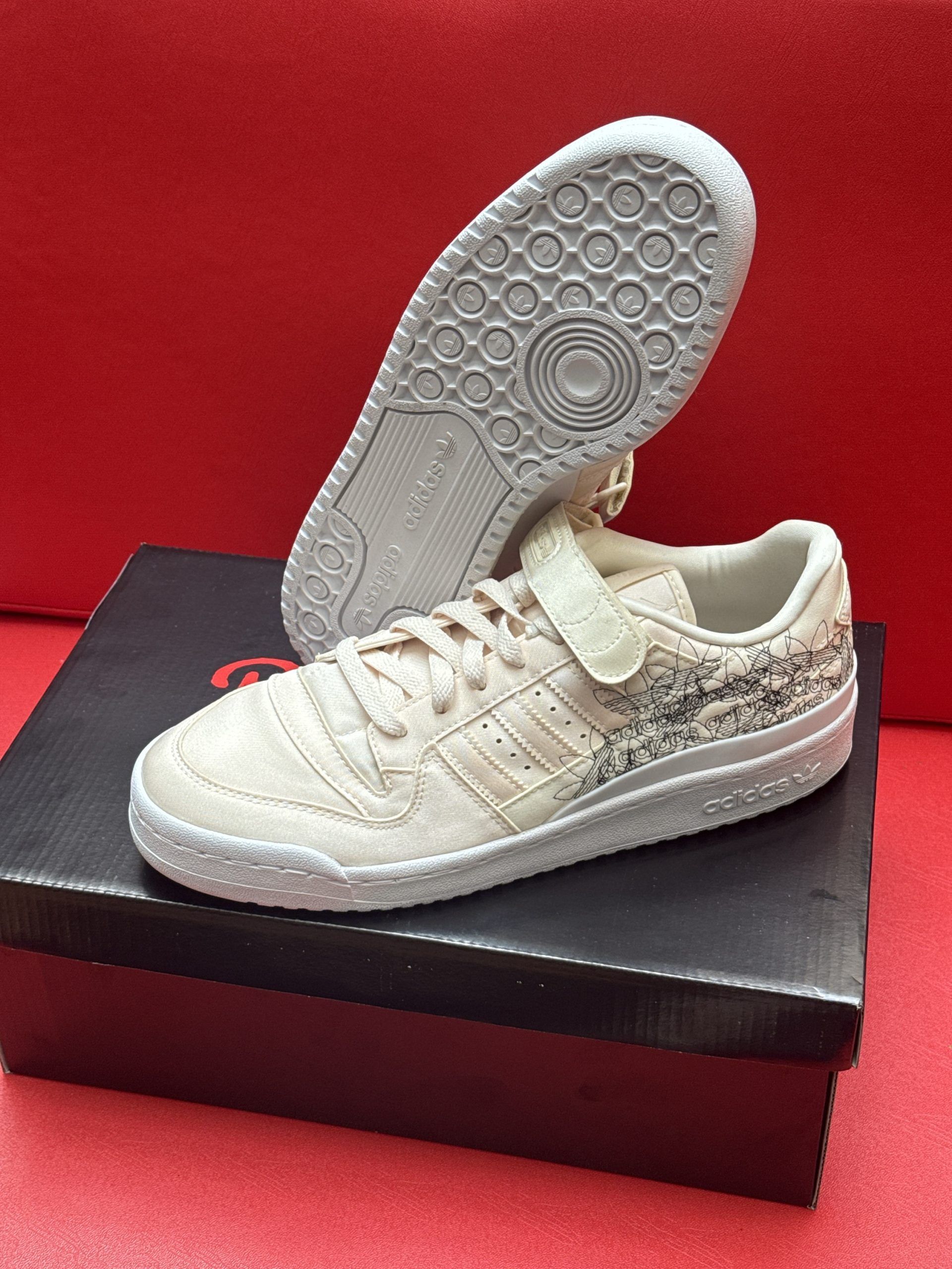 Pair of beige Adidas Forum Low sneakers on a black box, one shoe sole up, on a red surface.
