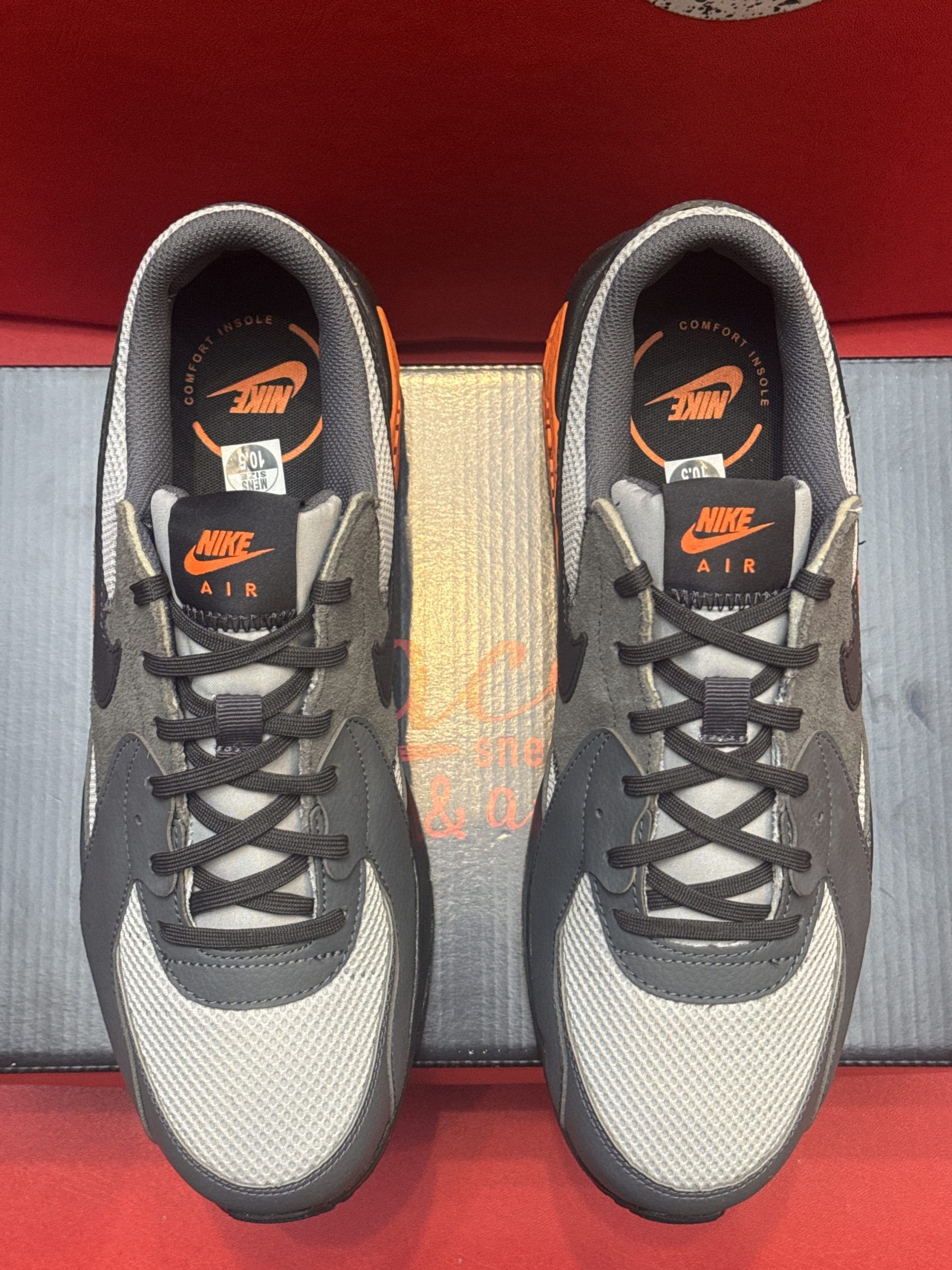 Gray, orange, and black Nike Air Max 90 sneakers on a black box, with a red background.
