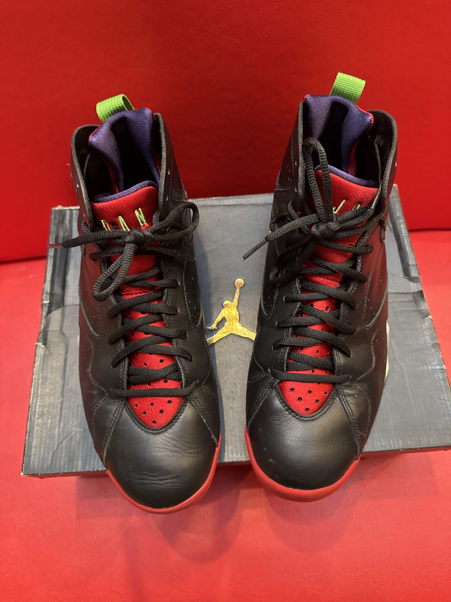 Pair of black, red, and green Air Jordan 7 sneakers on a box, red background.