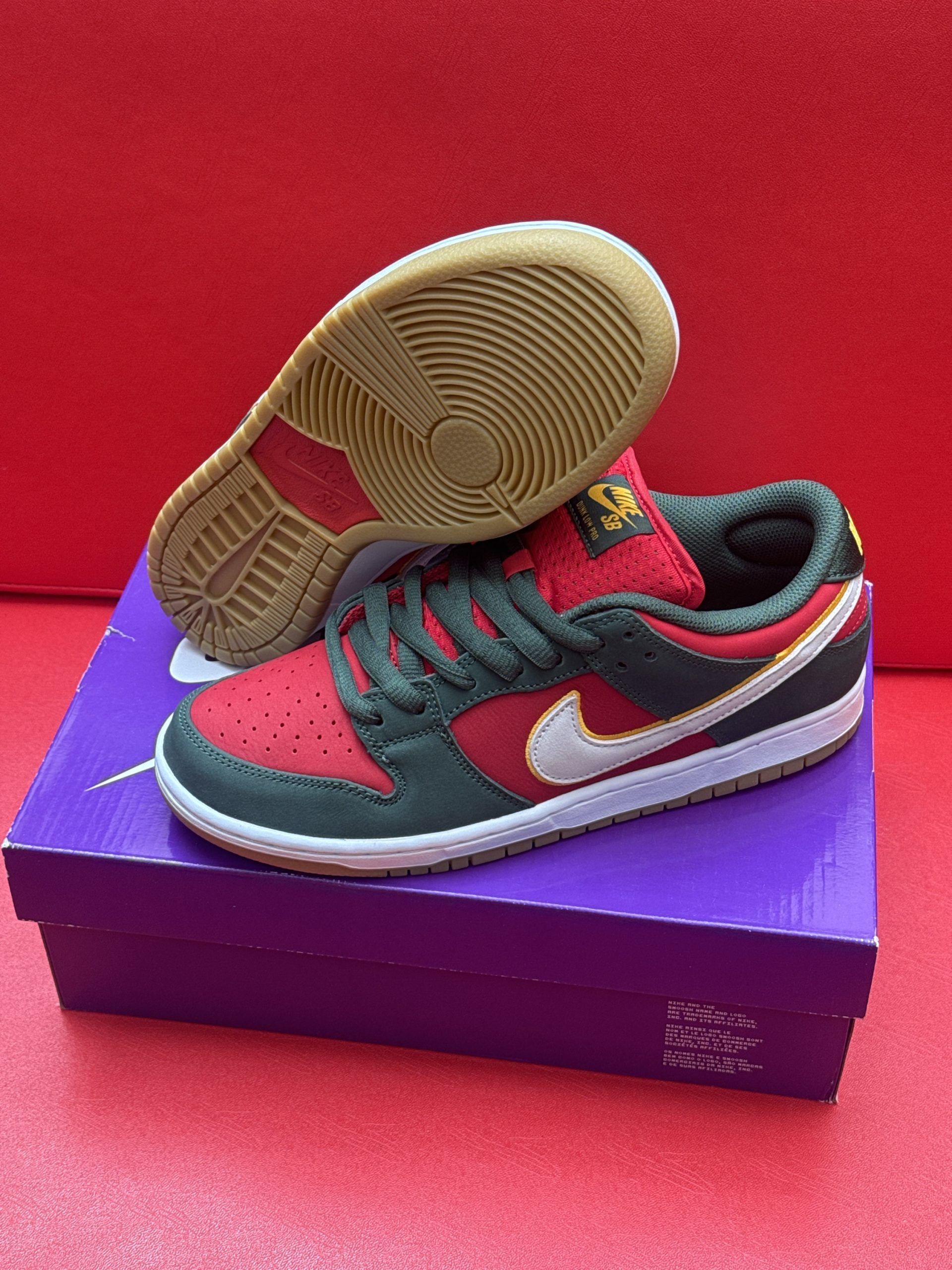 Nike SB Dunk Low sneakers, red, black, and white, on top of a purple shoebox with a brown sole on a red surface.