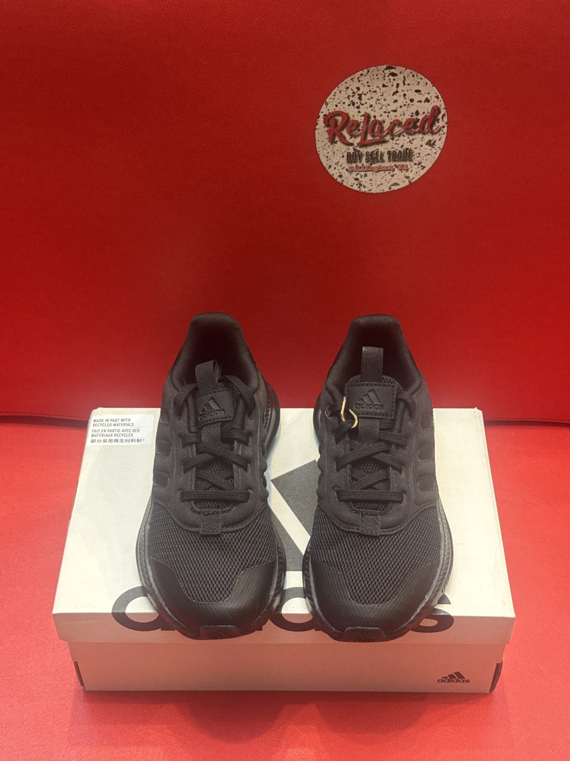 Black Adidas baby shoes on a white Adidas box, set against a red background.