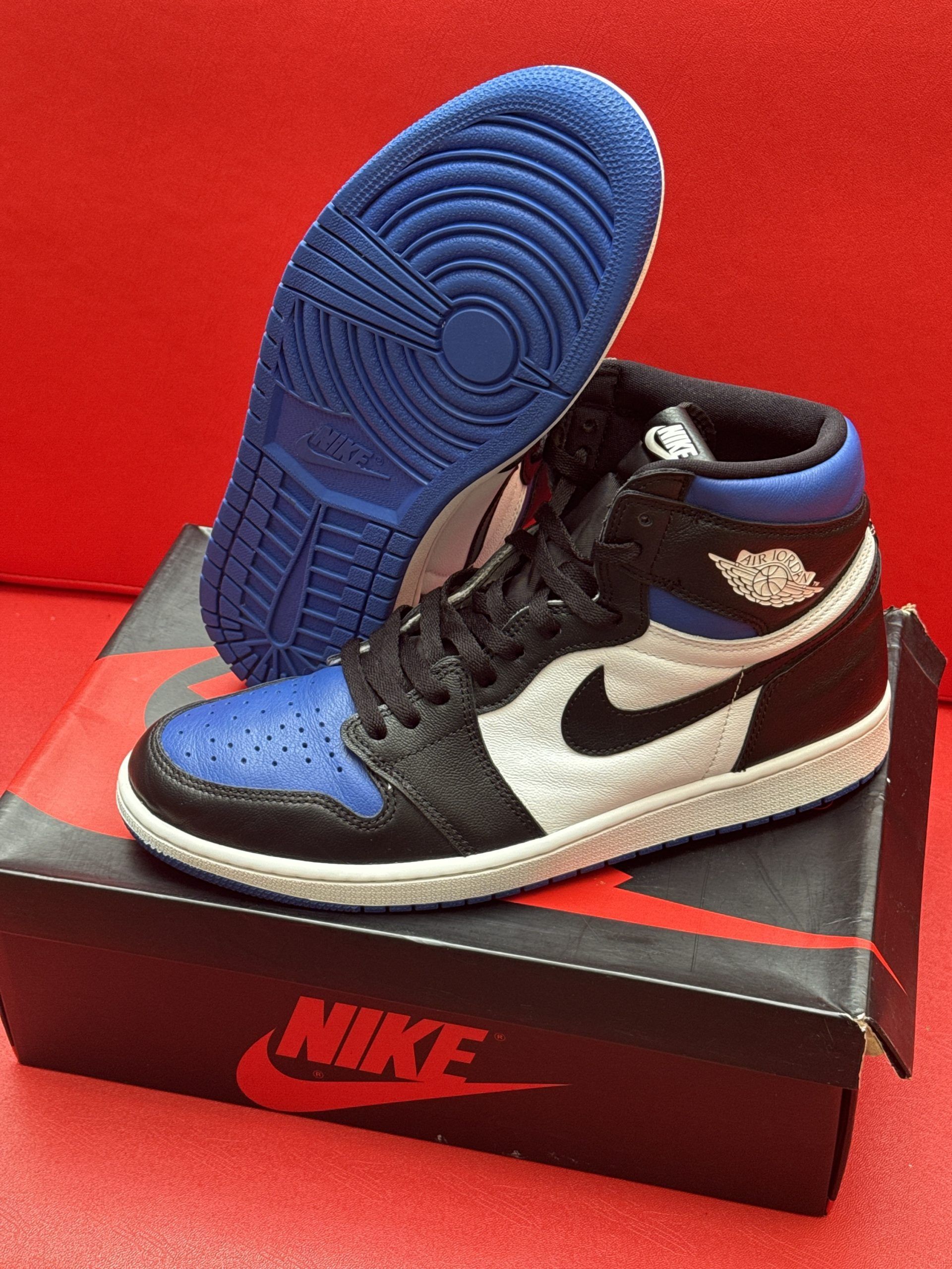 Pair of blue, black, and white Air Jordan 1 sneakers on a box with a red Nike logo; one shoe's sole is visible.