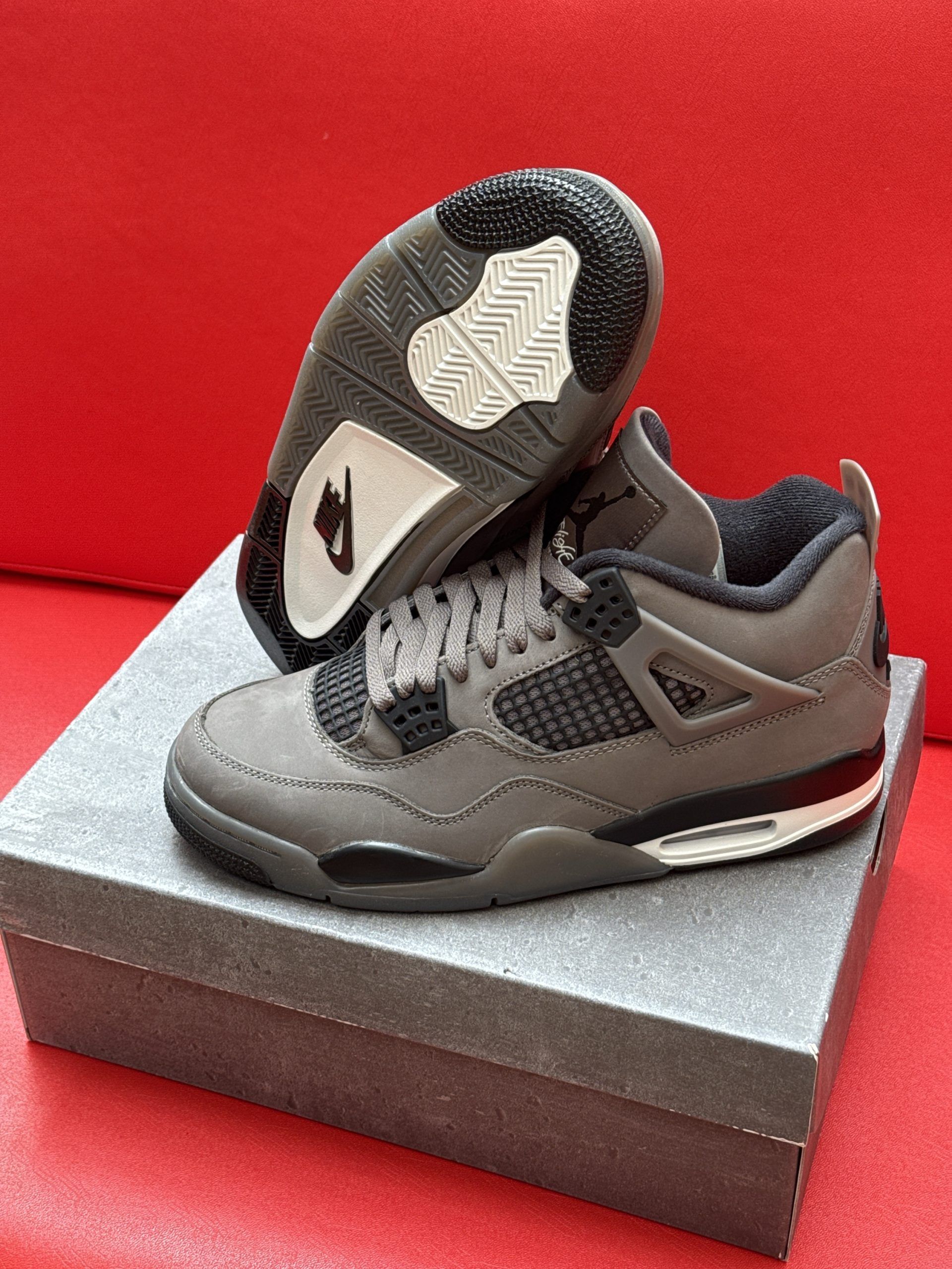 Gray and black Air Jordan 4 sneakers on a gray box, with the sole visible. Red background.