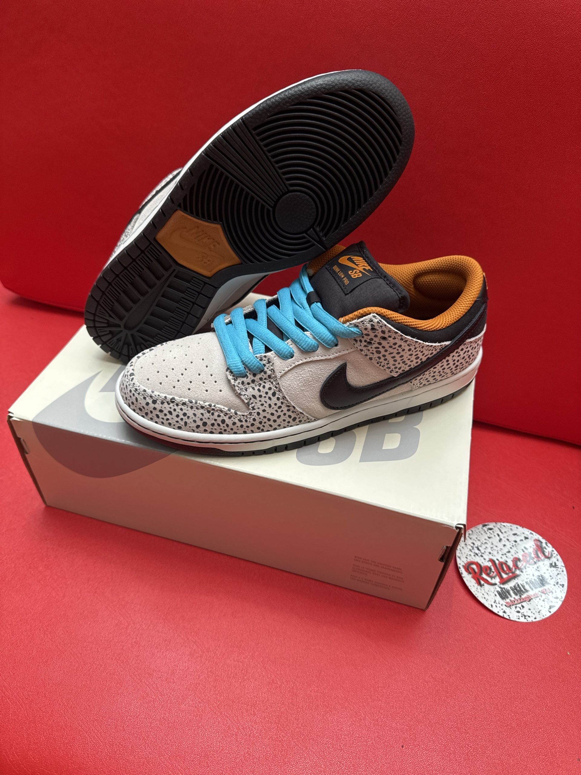 Nike SB Dunk Low sneakers, tan with black speckled pattern, blue laces, and orange collar, on a white box.