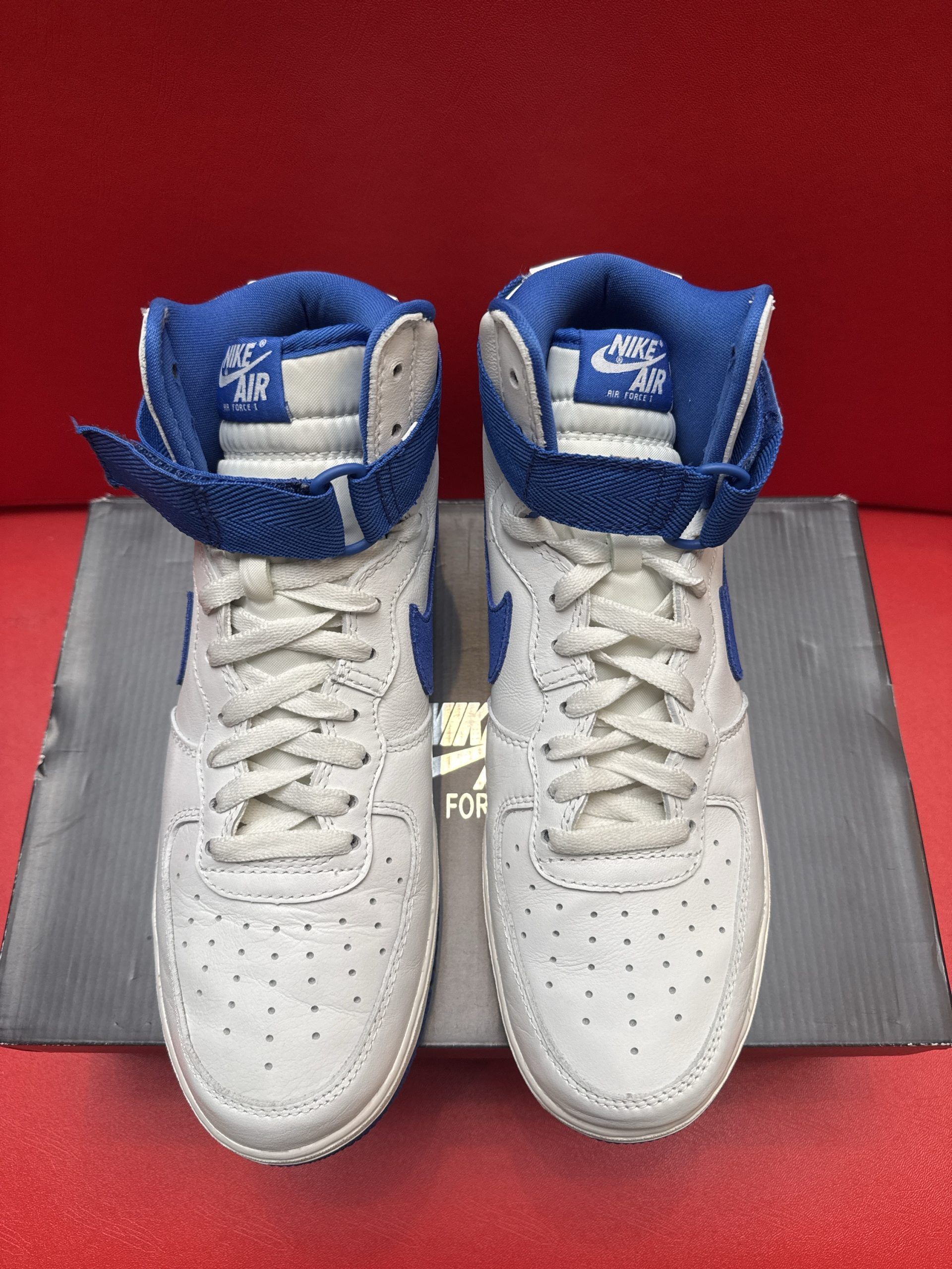 Pair of white and blue Nike high-top sneakers on a black shoebox, set against a red background.