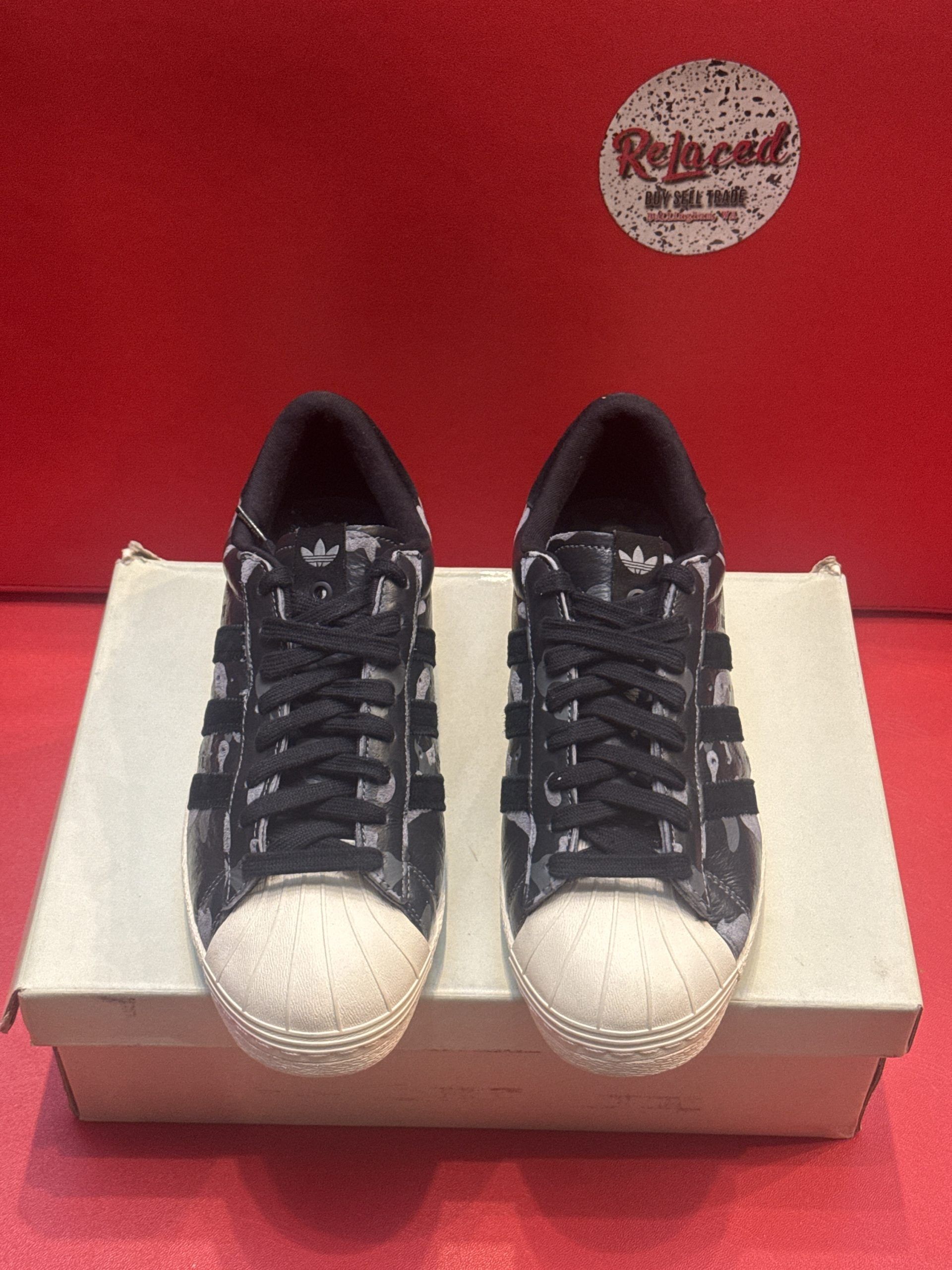 Adidas Superstar sneakers in a box. One sole is up, the other on the box, both white. Black and gray patterns on the shoes.