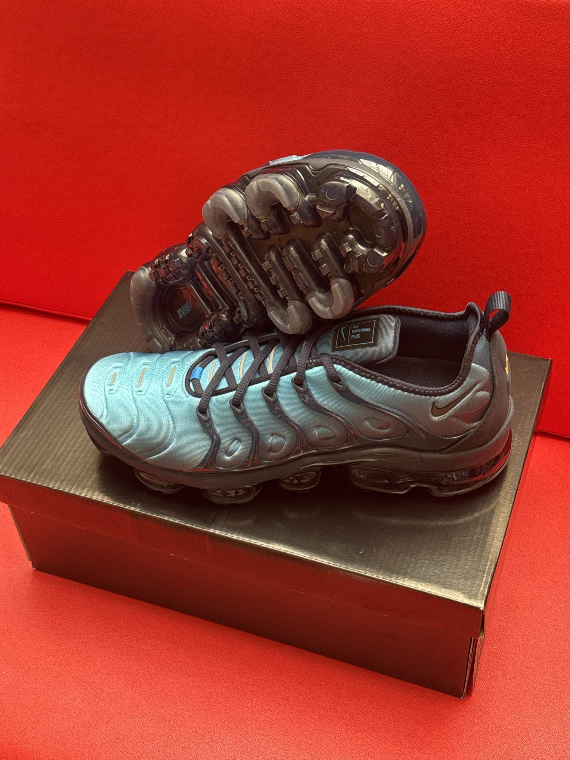 Blue and black Nike Air VaporMax Plus sneakers on a black shoebox, against a red backdrop.