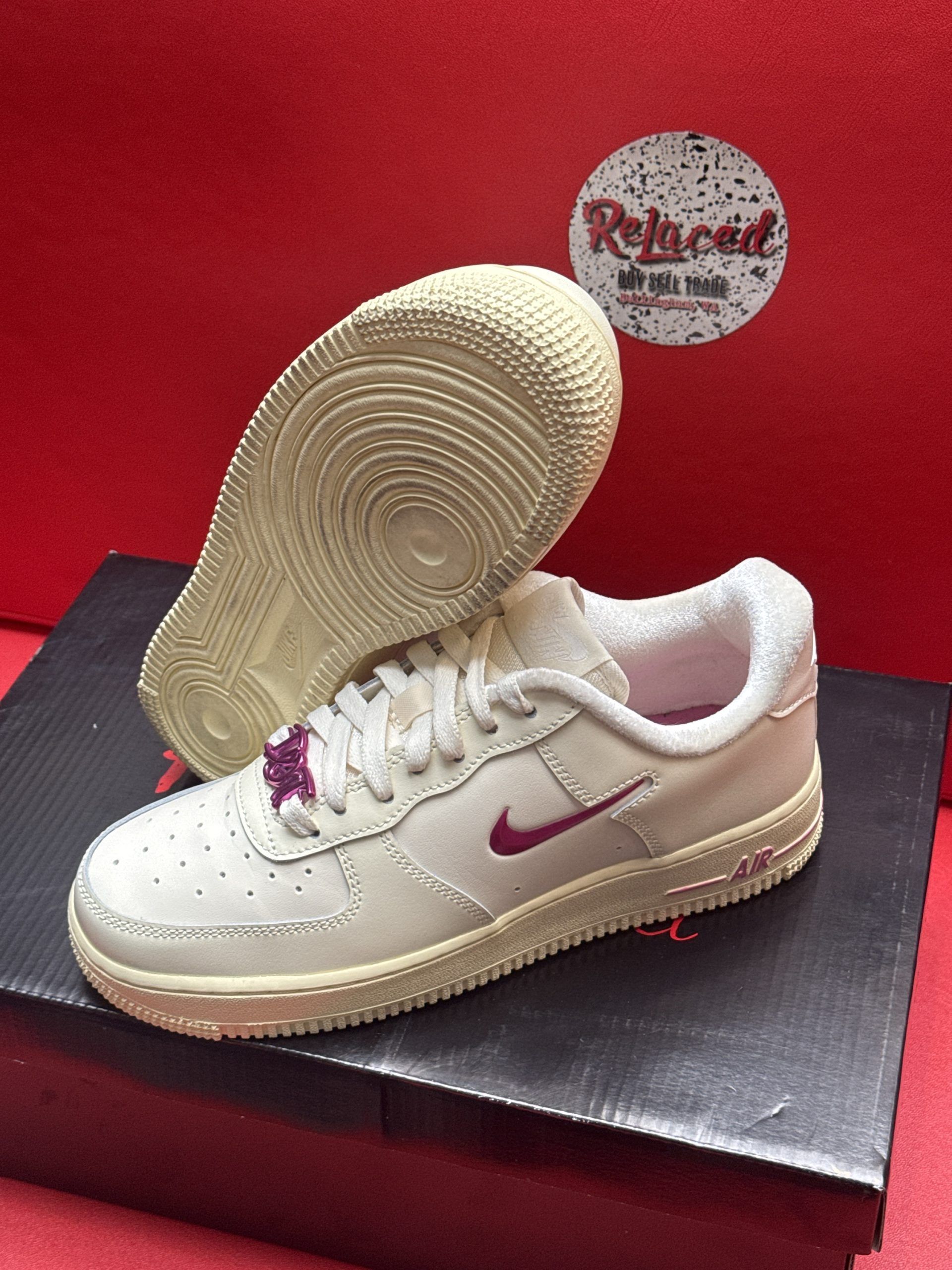 Cream-colored Nike Air Force 1 sneakers with a purple swoosh and laces, set on a dark box against a red backdrop.