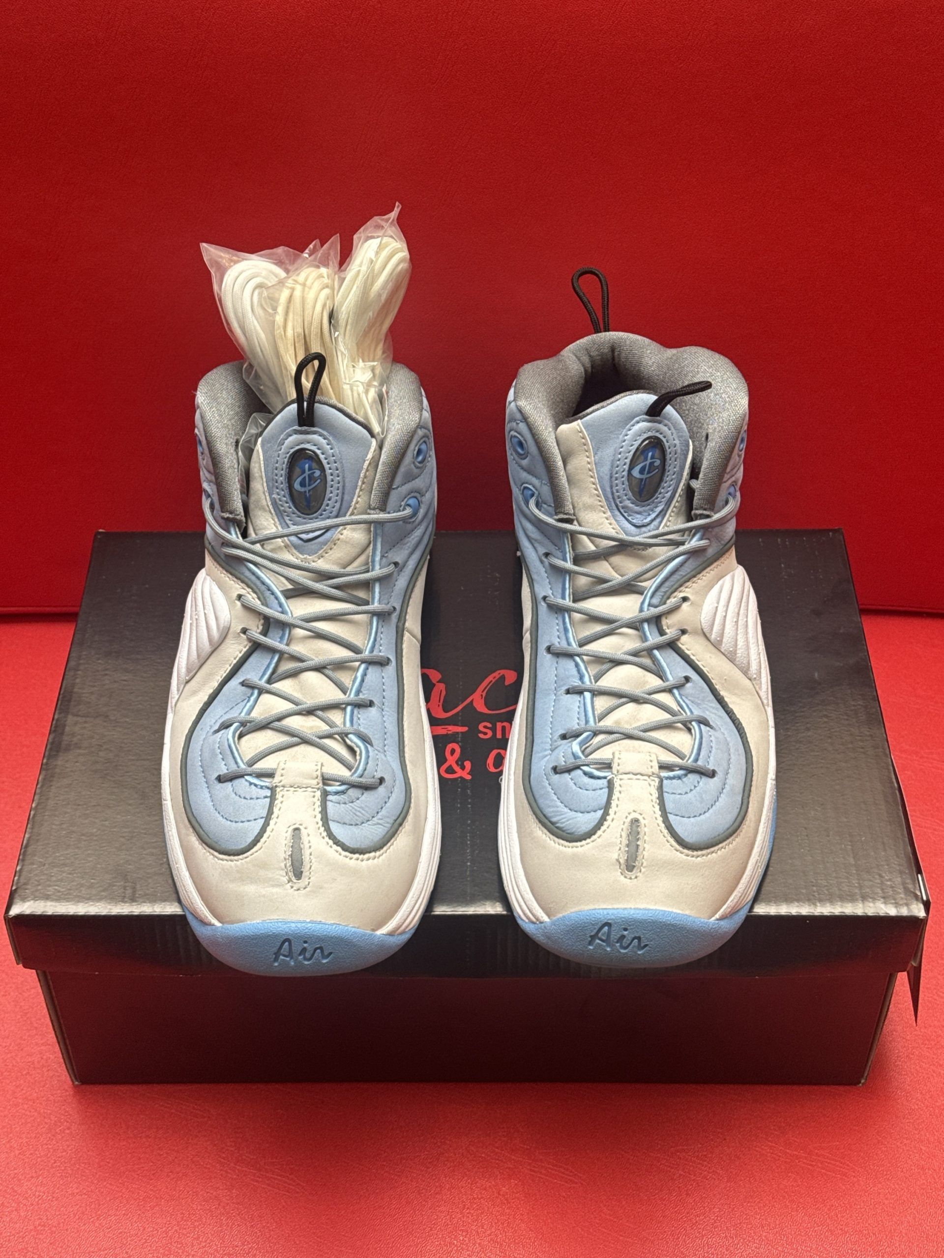 Light blue and gray Nike Penny Hardaway sneakers on a black box, red background.