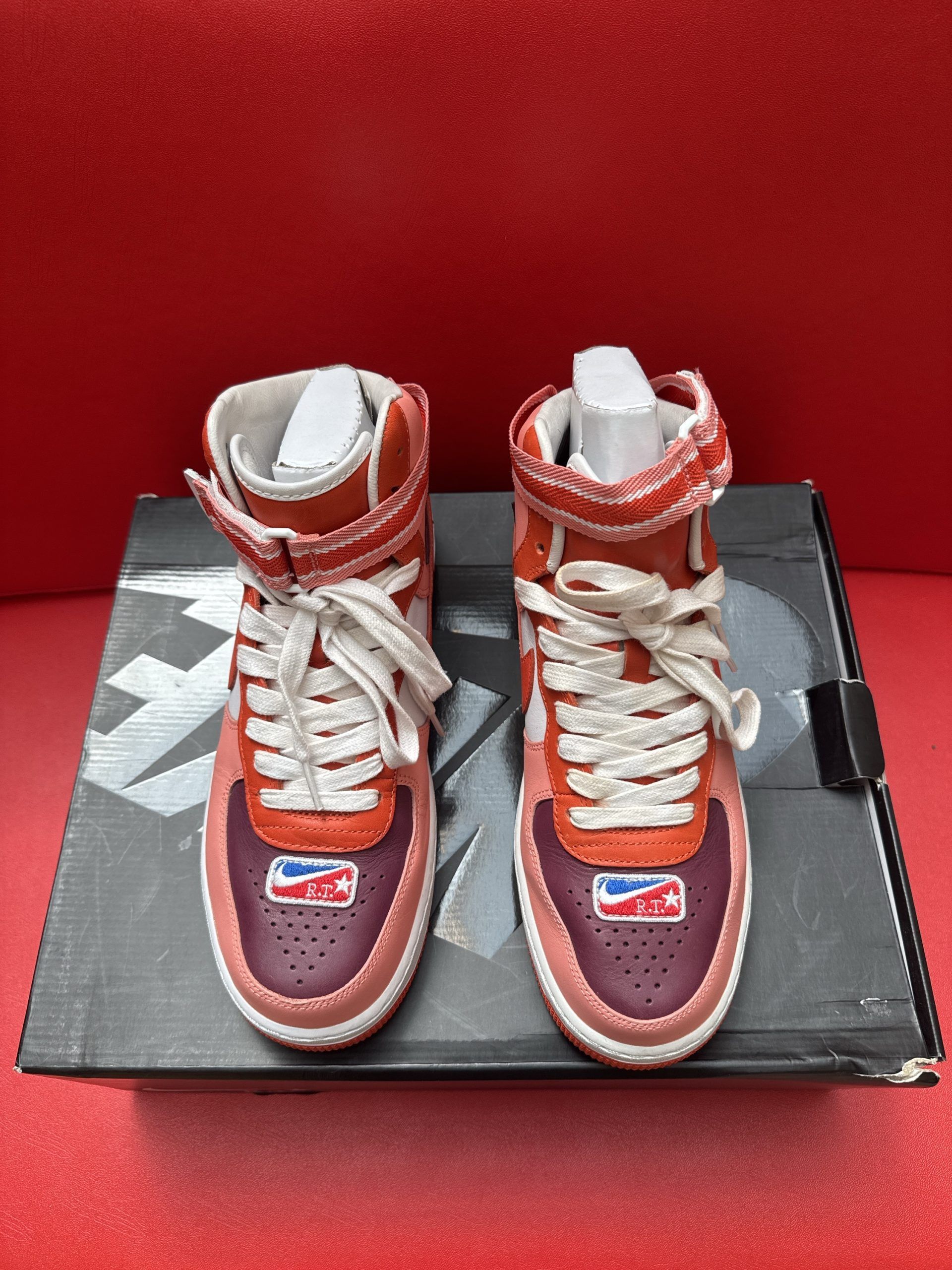 Red and pink Nike Air Force 1 high-top sneaker on top of a black Nike shoe box with a red sole.