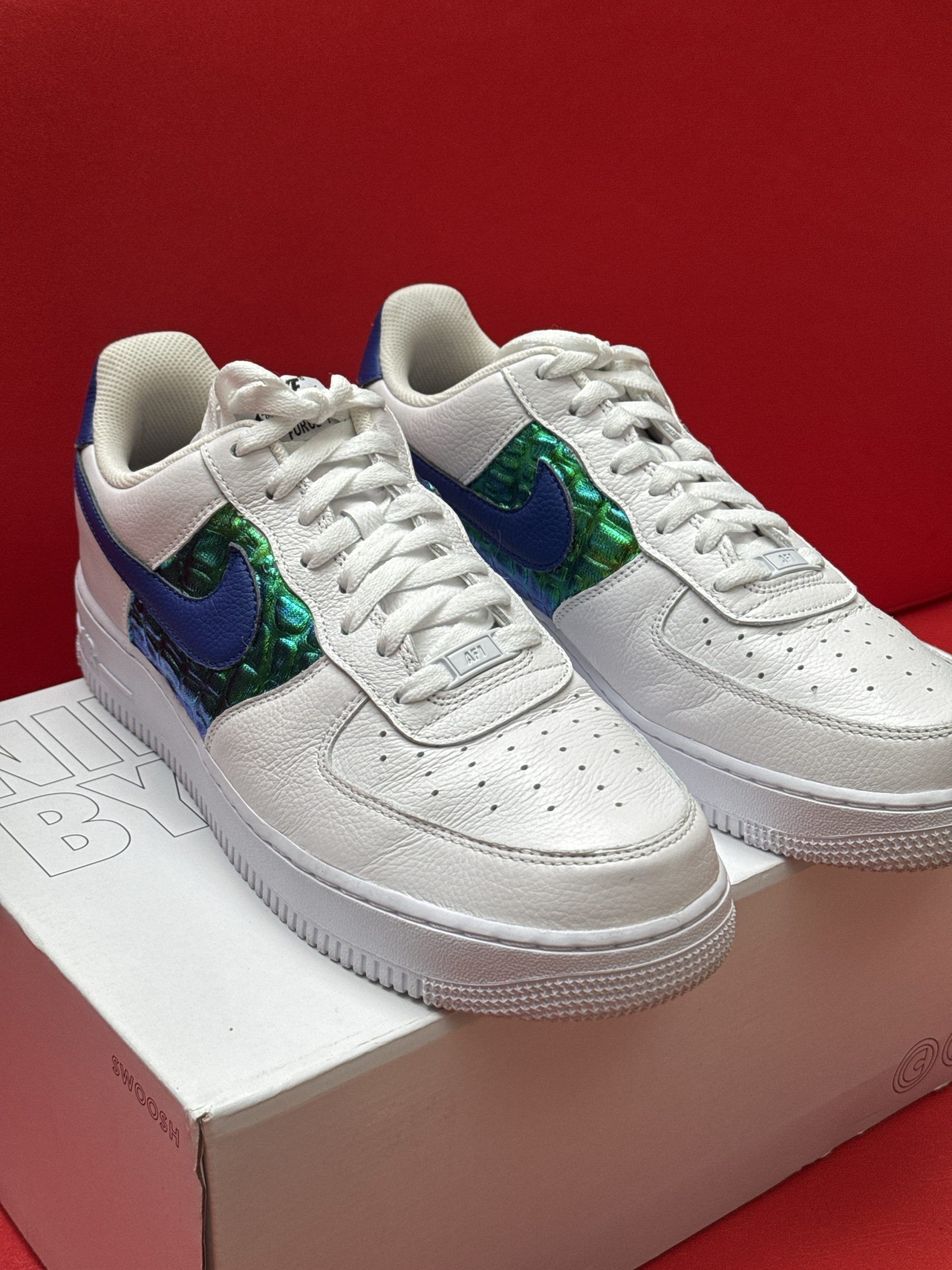 White Nike Air Force 1 sneakers with blue and green accents sit atop a white box against a red background.