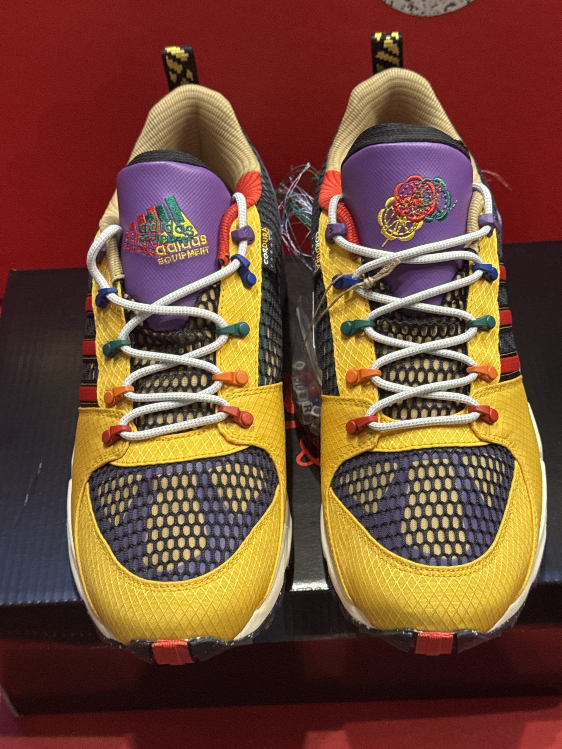 Adidas sneakers, yellow and purple, on a box, with a red background.