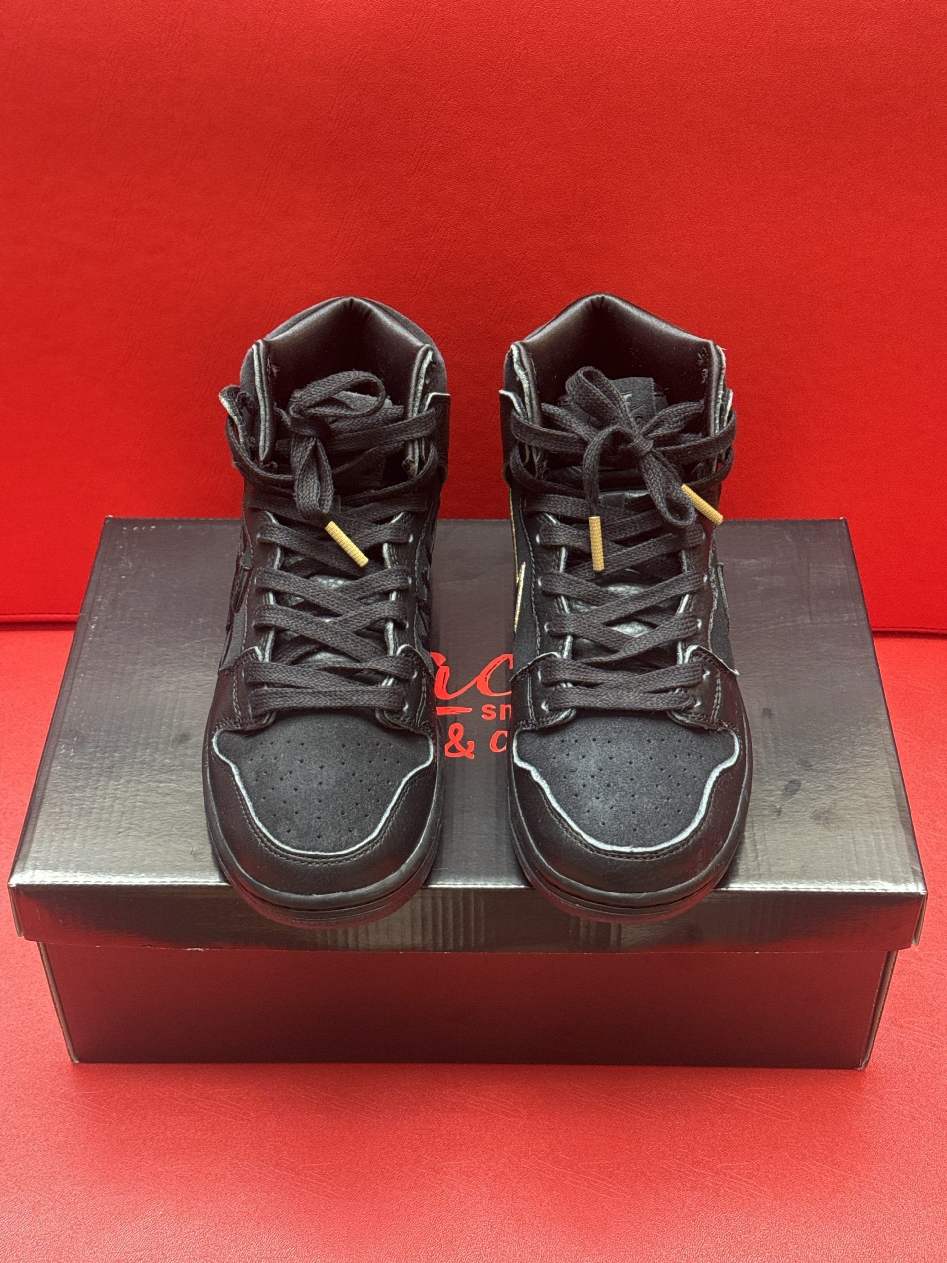 Black Nike SB Dunk High sneakers with gold accents, on a black shoebox.