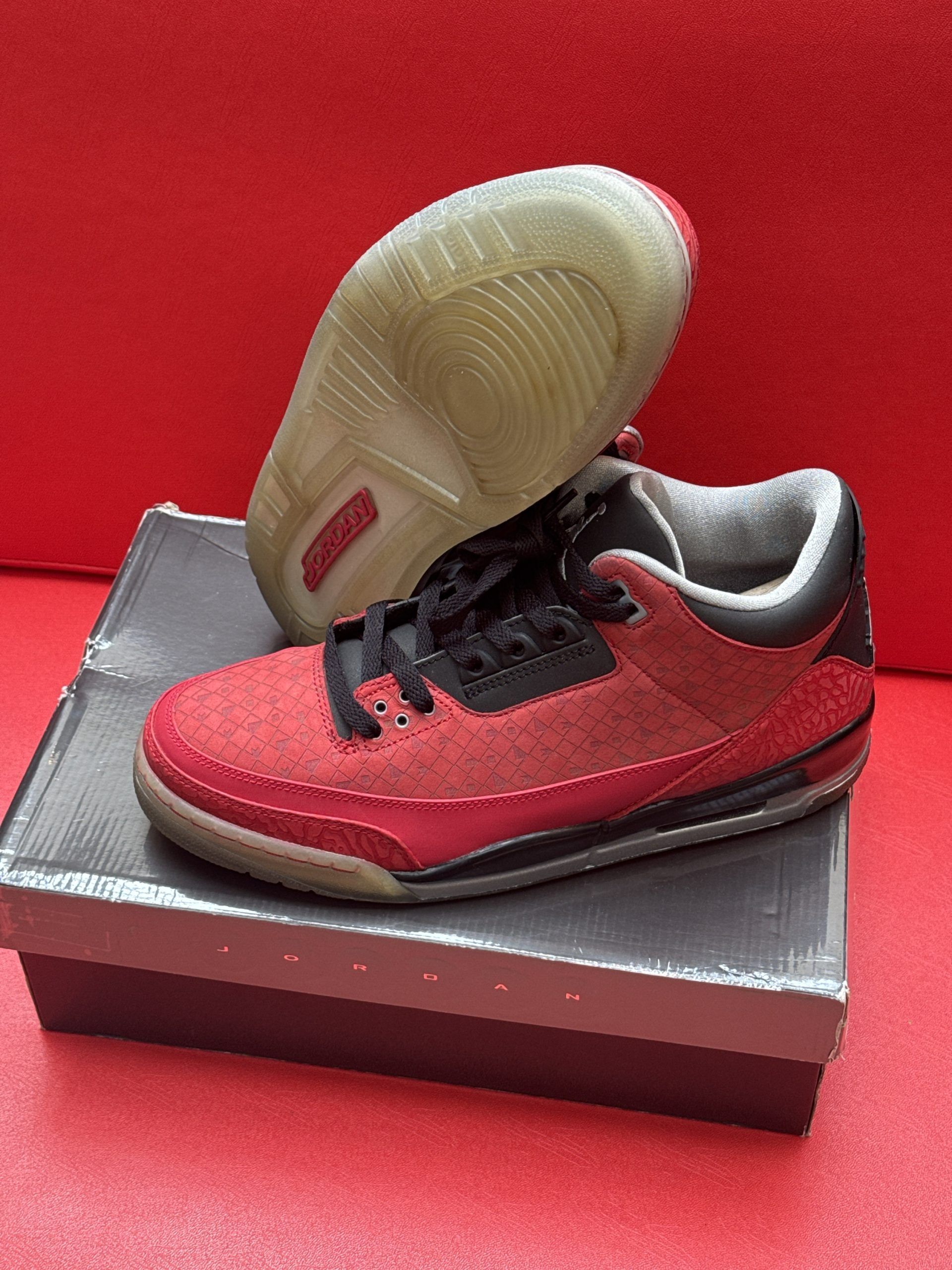 Red and black Air Jordan 3 sneakers with a worn sole on a shoebox against a red background.