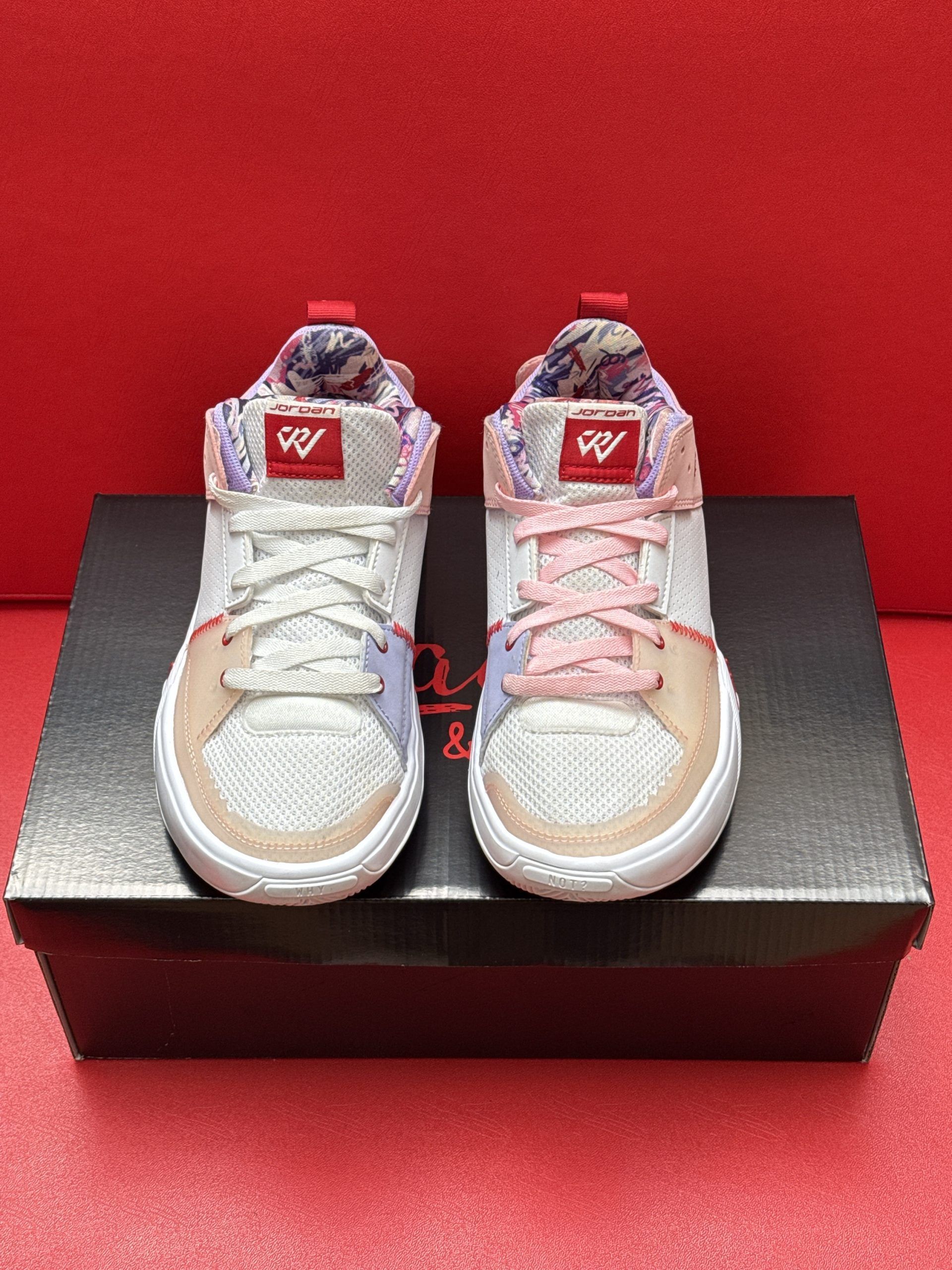 White, pink, and red Jordan sneakers on a black shoe box; red background.