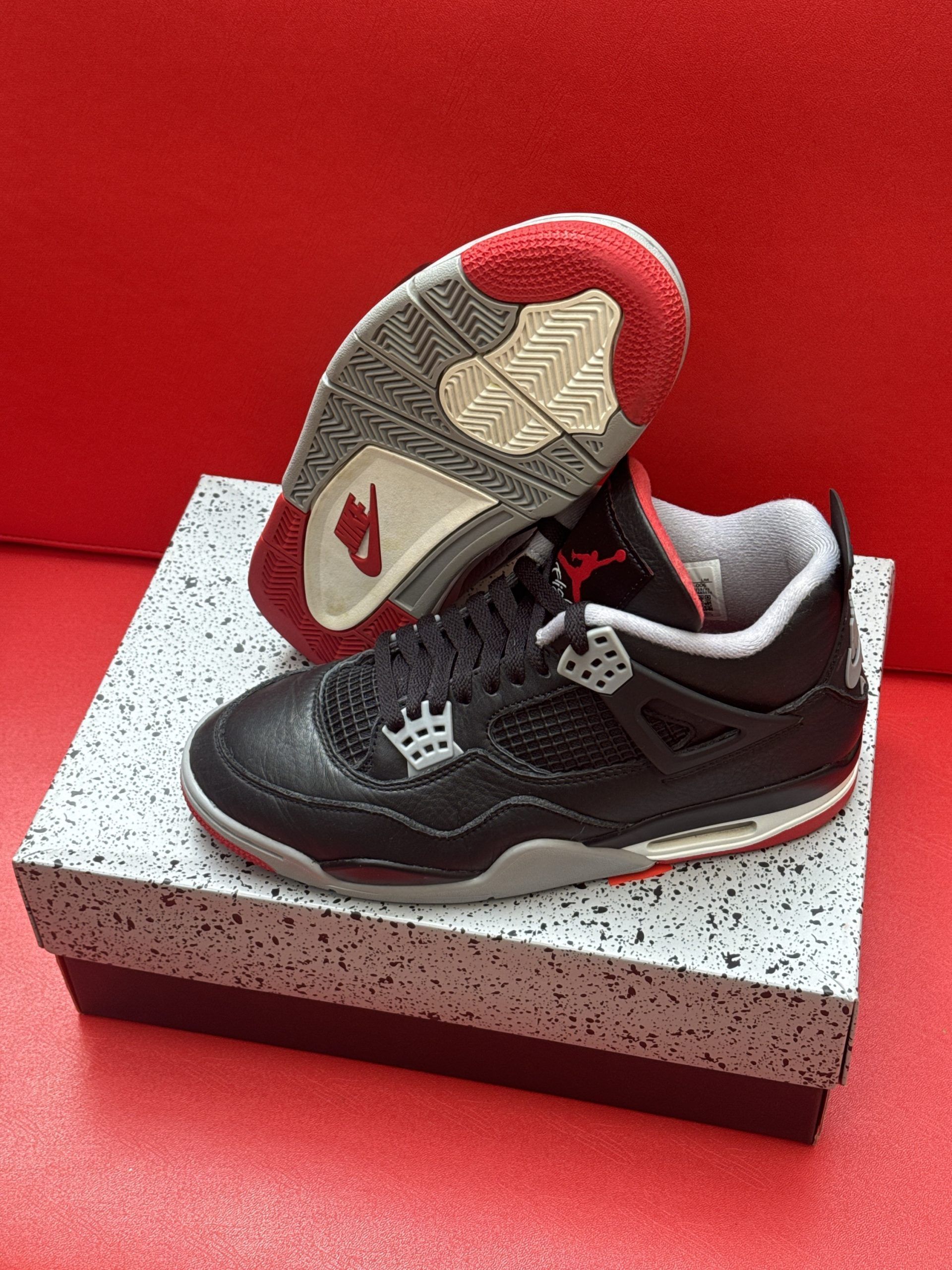 Black, red, and gray Air Jordan 4 infant sneakers on a speckled shoebox. One sneaker's sole is visible.