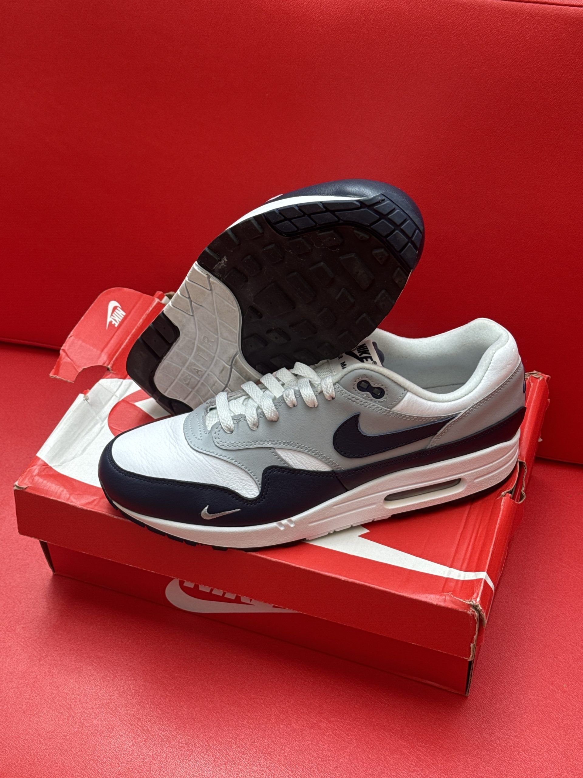 Nike Air Max 1 sneakers in gray, navy, and white on a red box.