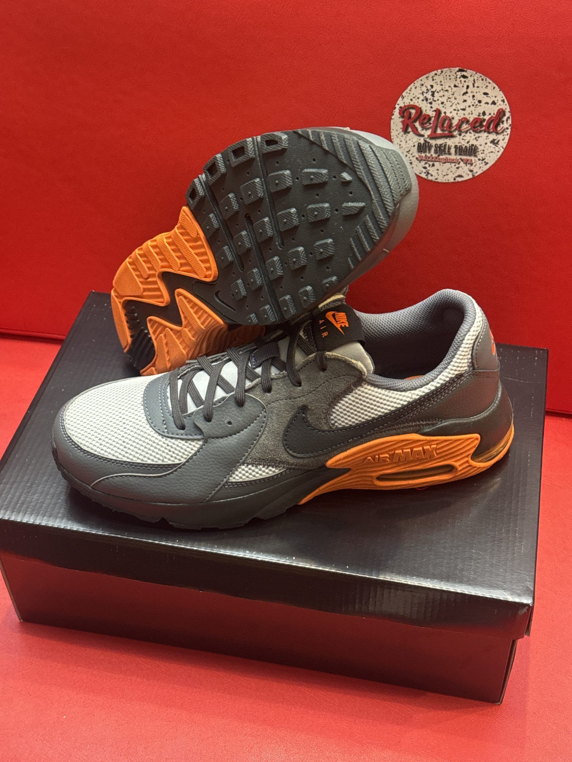 Gray, orange, and black Nike Air Max 90 sneakers on a black box, with a red background.