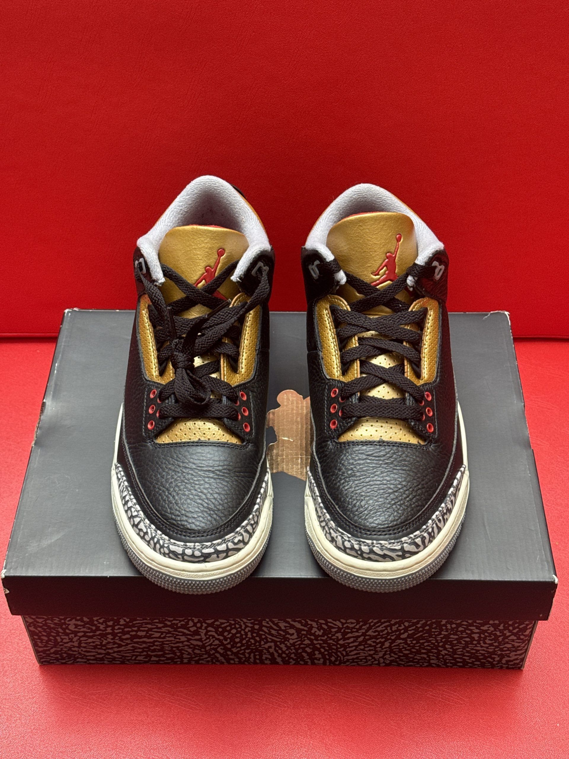 Black, gold, and red Air Jordan 3 sneakers on a black box, sole visible, set against a red background.
