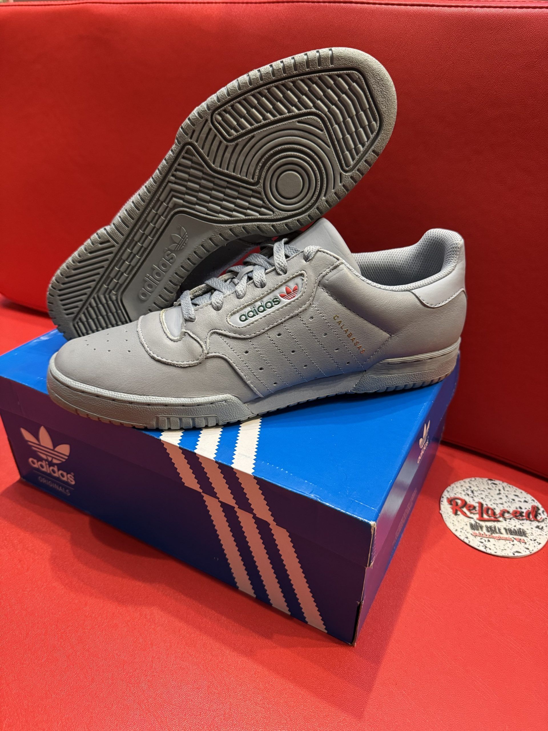 Gray Adidas sneakers on blue box, red background. Sole up.