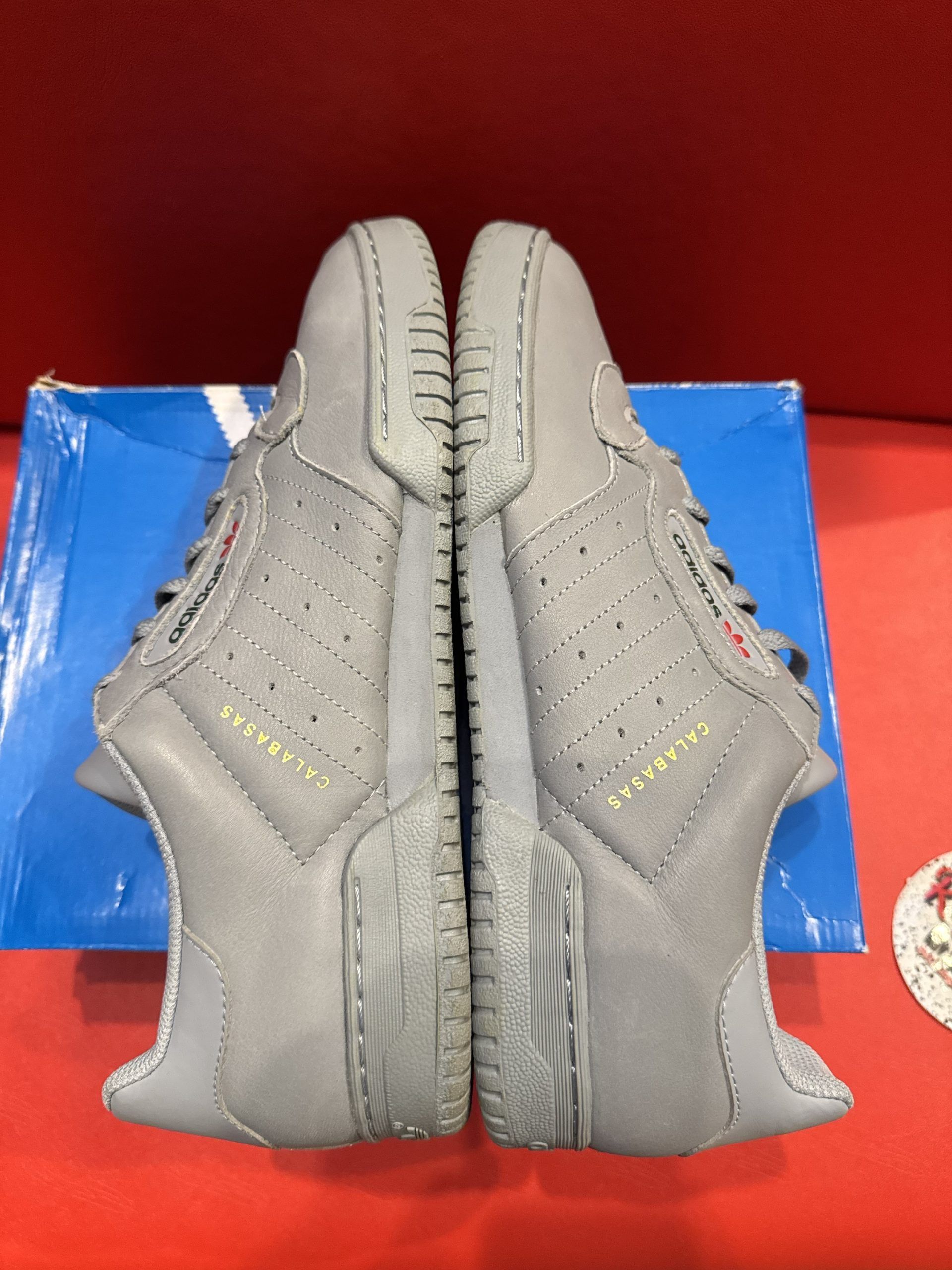 Gray Adidas sneakers on blue box, red background. Sole up.