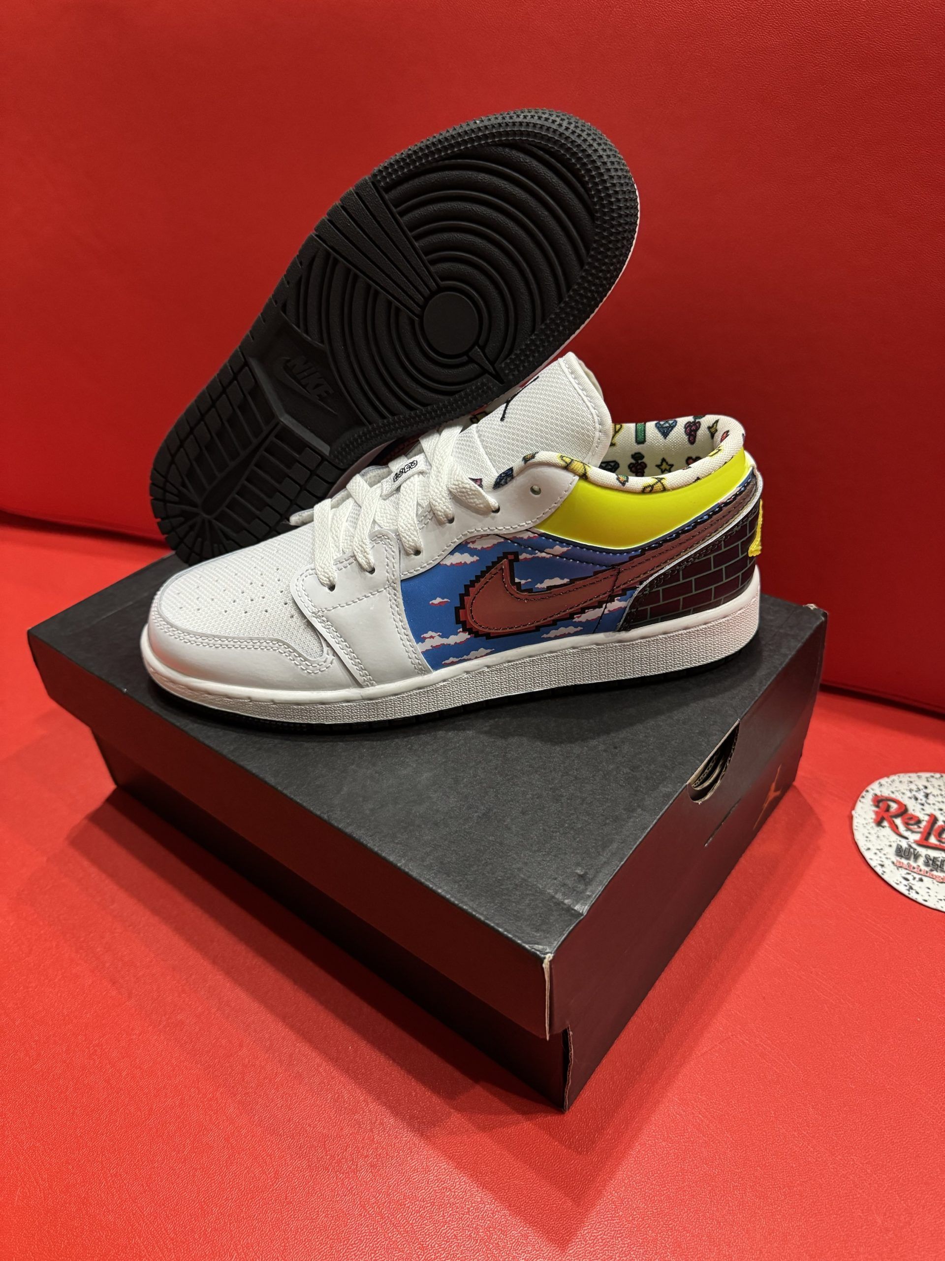 White and multi-colored Air Jordan 1 sneaker on a black shoebox, sole in background, set against a red background.