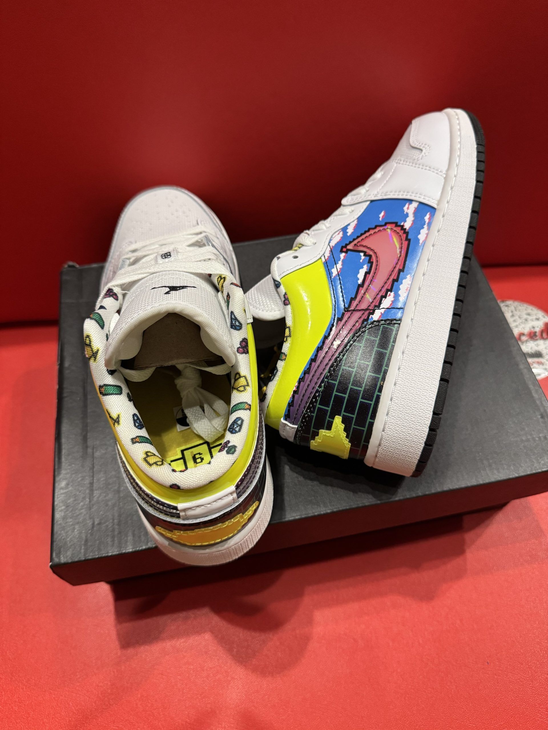 White and multi-colored Air Jordan 1 sneaker on a black shoebox, sole in background, set against a red background.