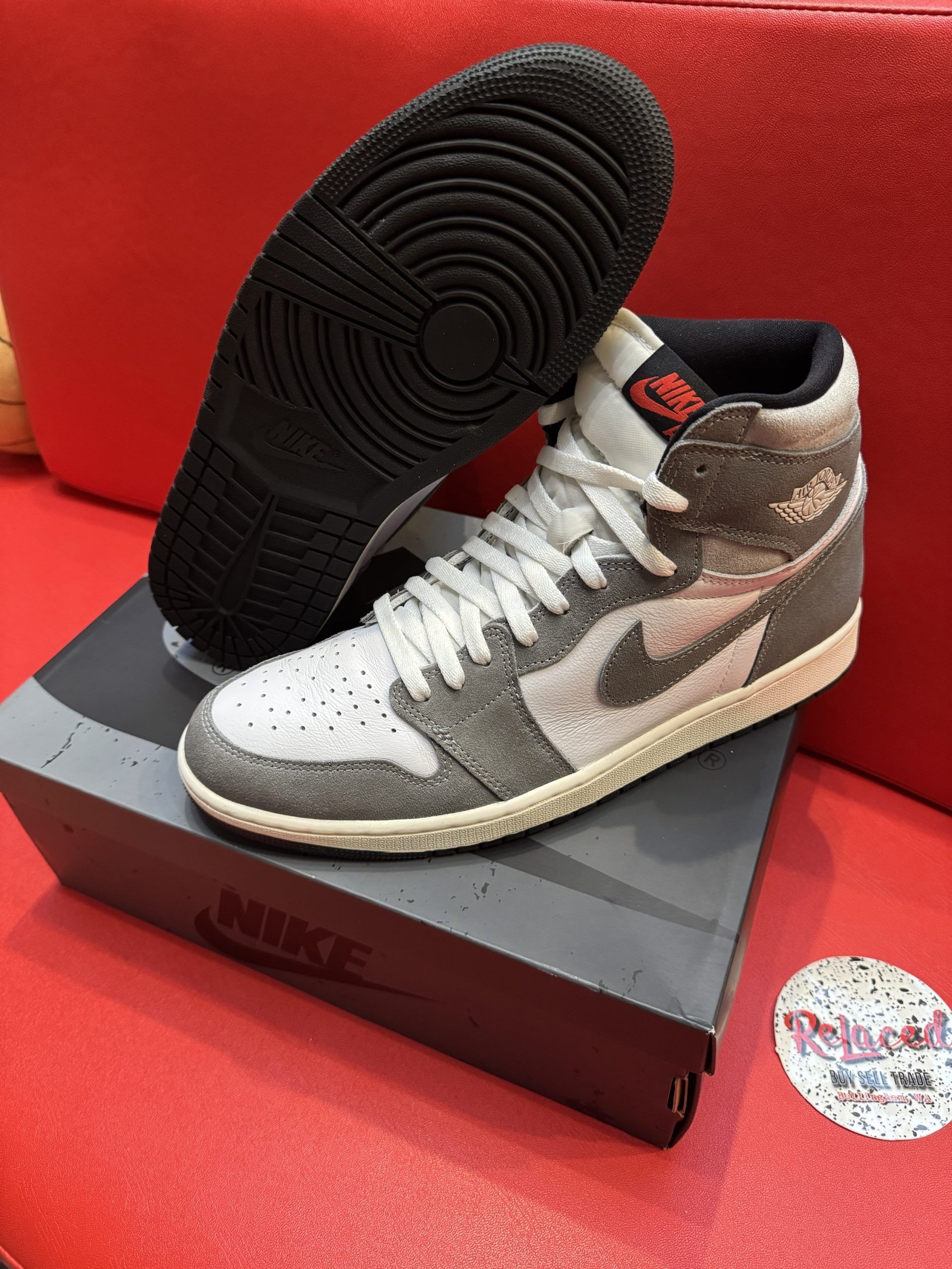 Air Jordan 1 high-top sneakers, gray and white with a black sole, resting on a gray box, on a red surface.