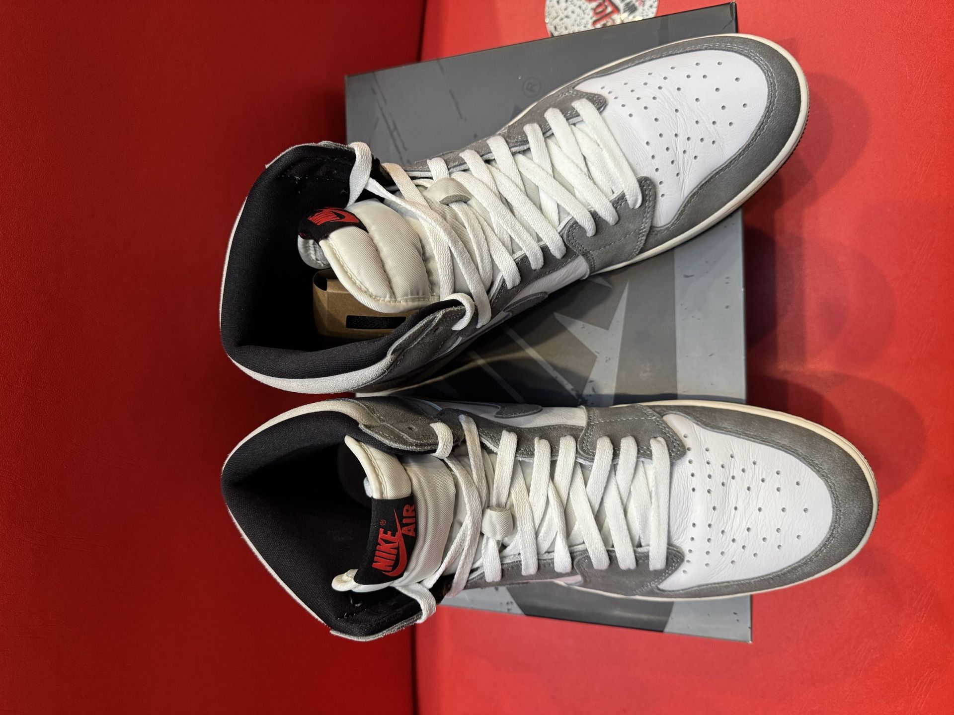 Air Jordan 1 high-top sneakers, gray and white with a black sole, resting on a gray box, on a red surface.