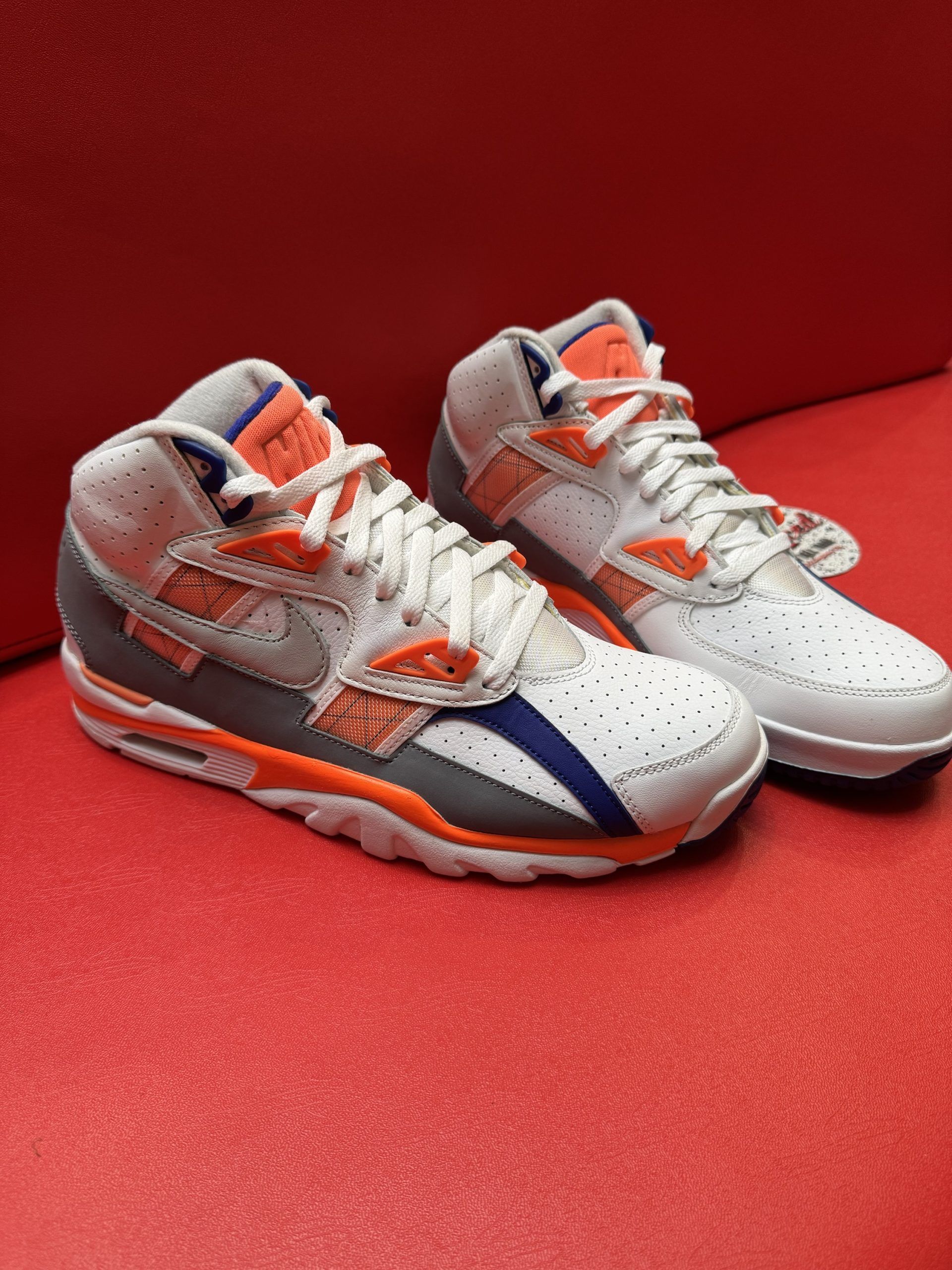 White Nike Air Trainer sneakers with orange and blue accents, on red surface.