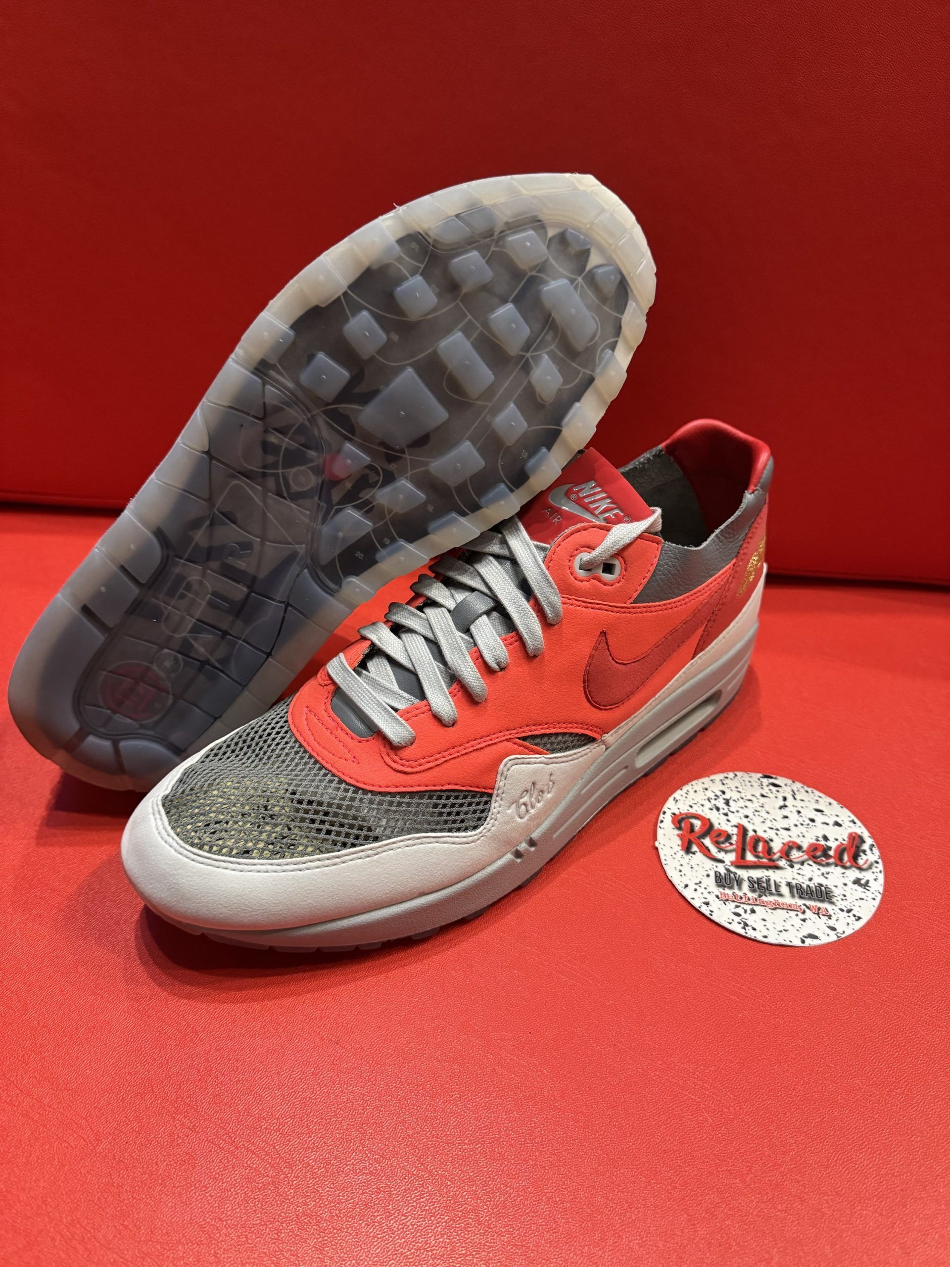 Red and grey Nike Air Max 1 sneaker with a transparent sole, sitting on a red surface.