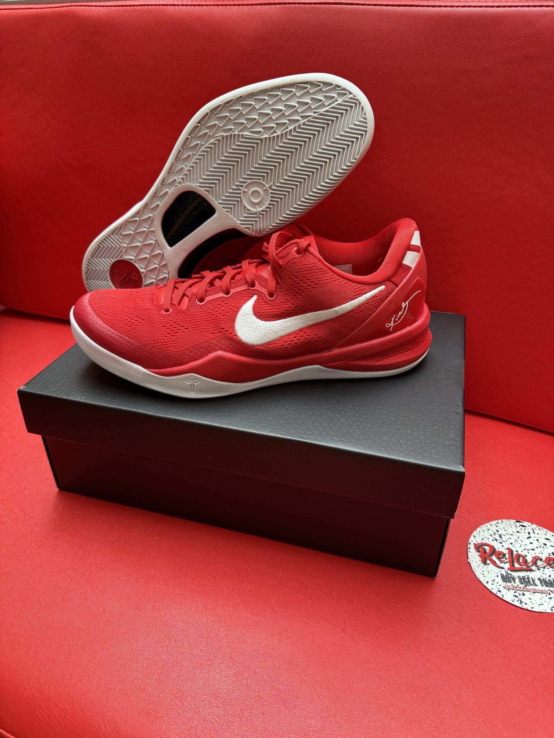 Red Nike basketball shoe with white swoosh, on a black box, with the sole visible. Red background.