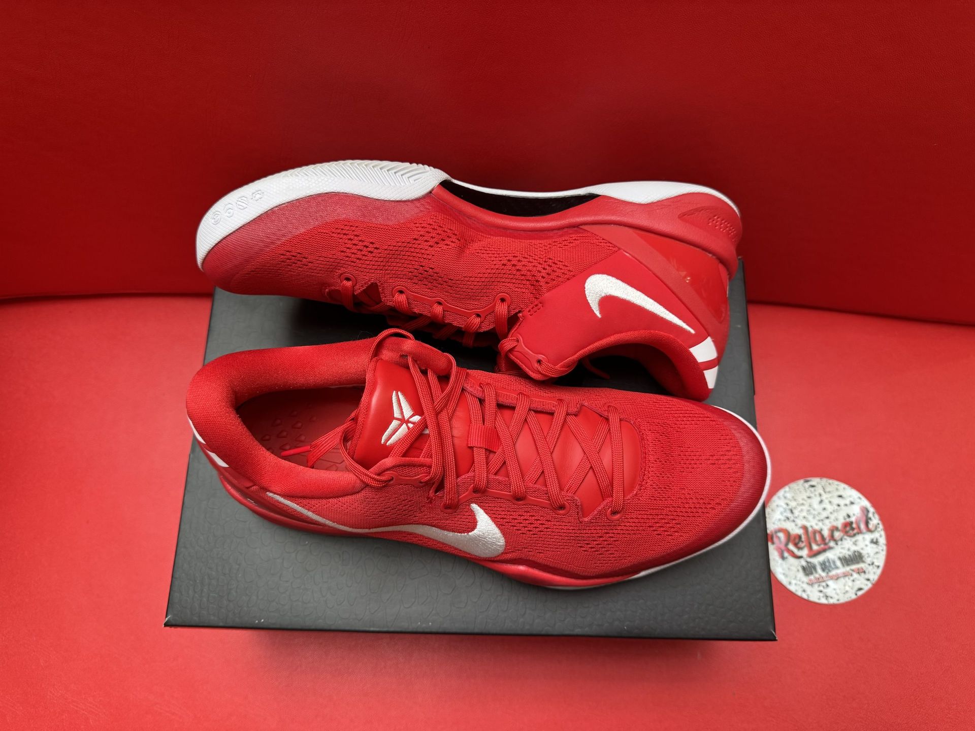 Red Nike basketball shoe with white swoosh, on a black box, with the sole visible. Red background.