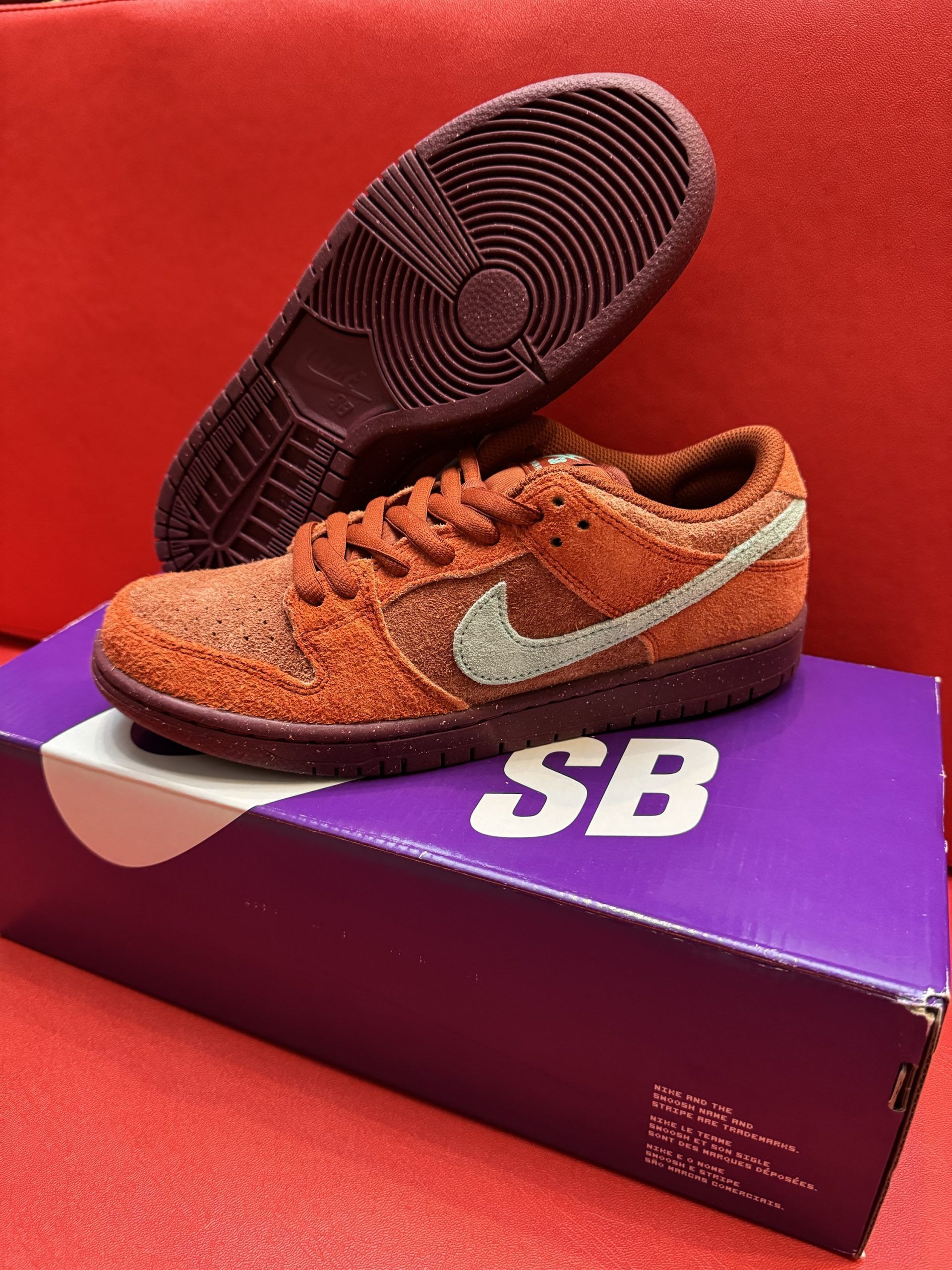 Two brown Nike Dunk SB sneakers on a purple Nike shoebox, sitting on a red surface.