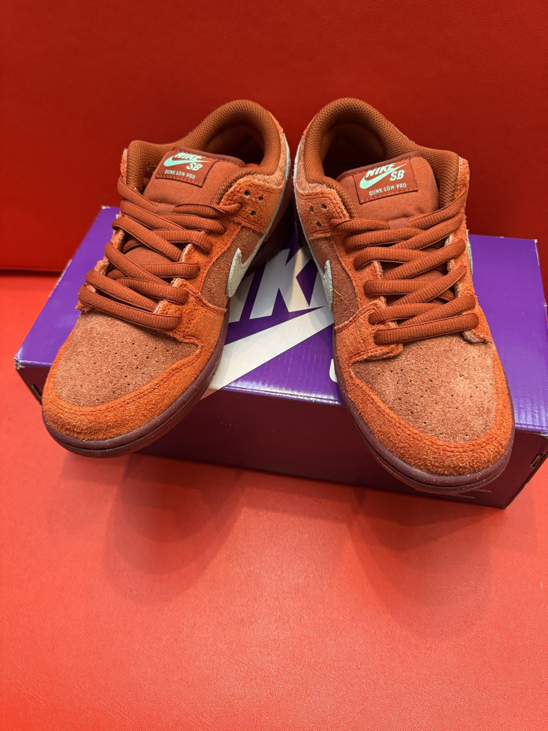 Two brown Nike Dunk SB sneakers on a purple Nike shoebox, sitting on a red surface.