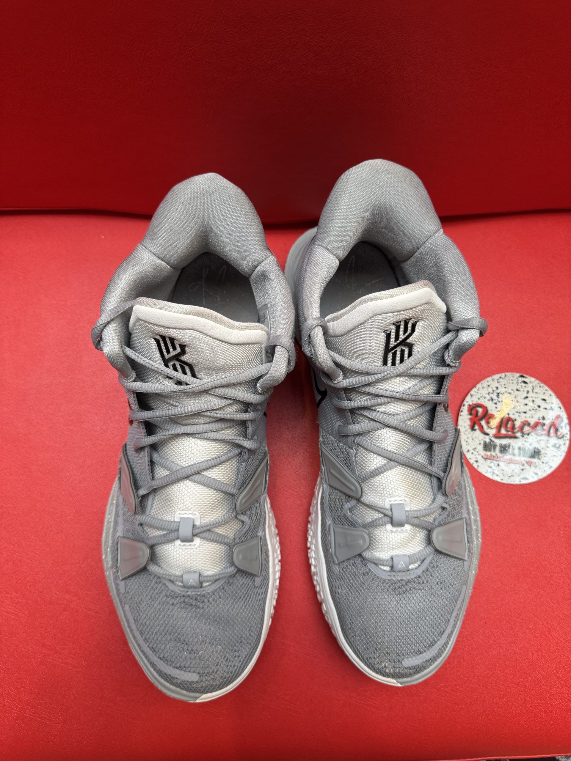 Gray and white Nike Kyrie sneakers on a red surface, sole facing up. Black swoosh.