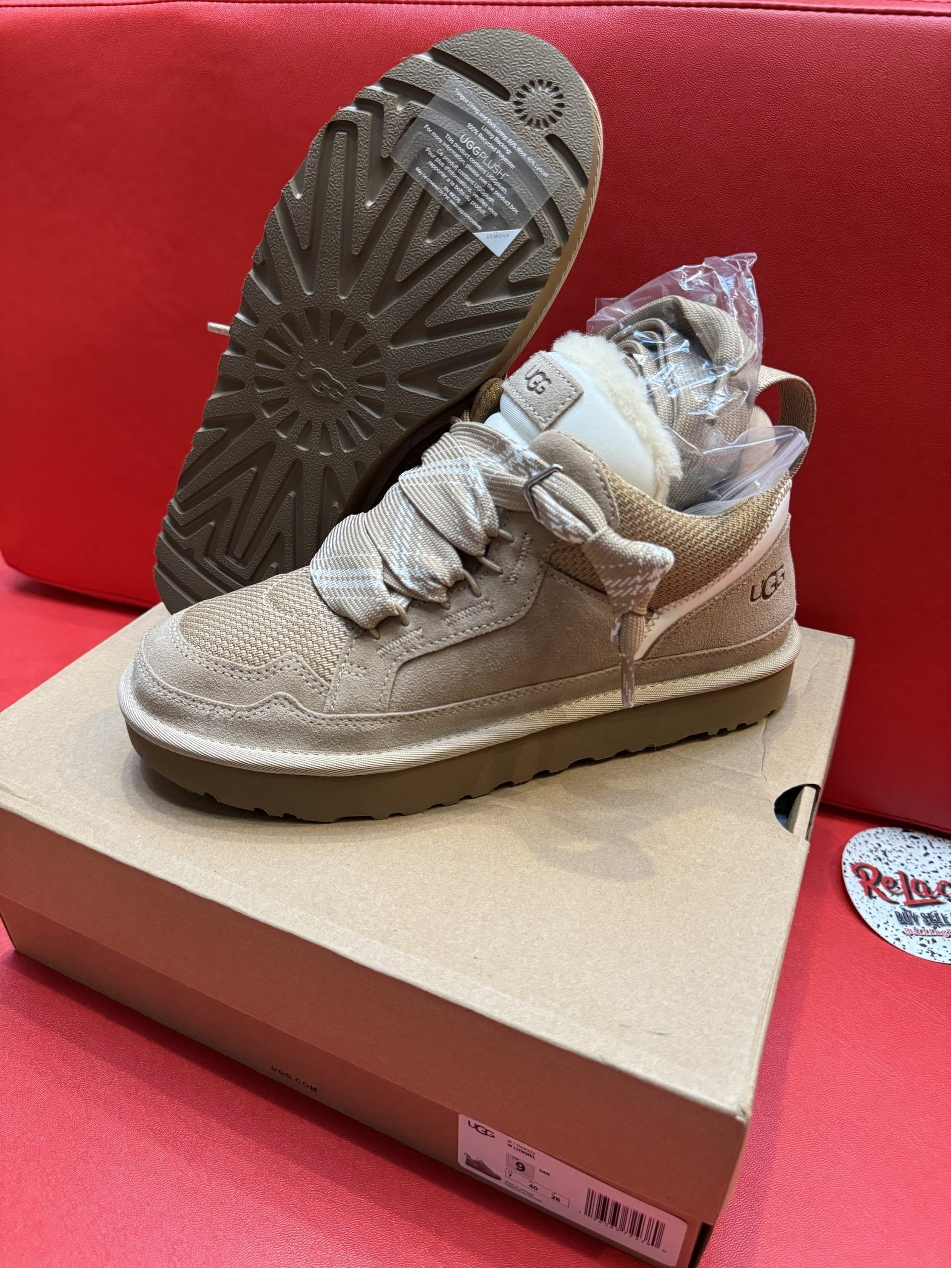 Tan UGG sneaker on a cardboard box with the sole of a matching shoe in the background, resting on a red surface.