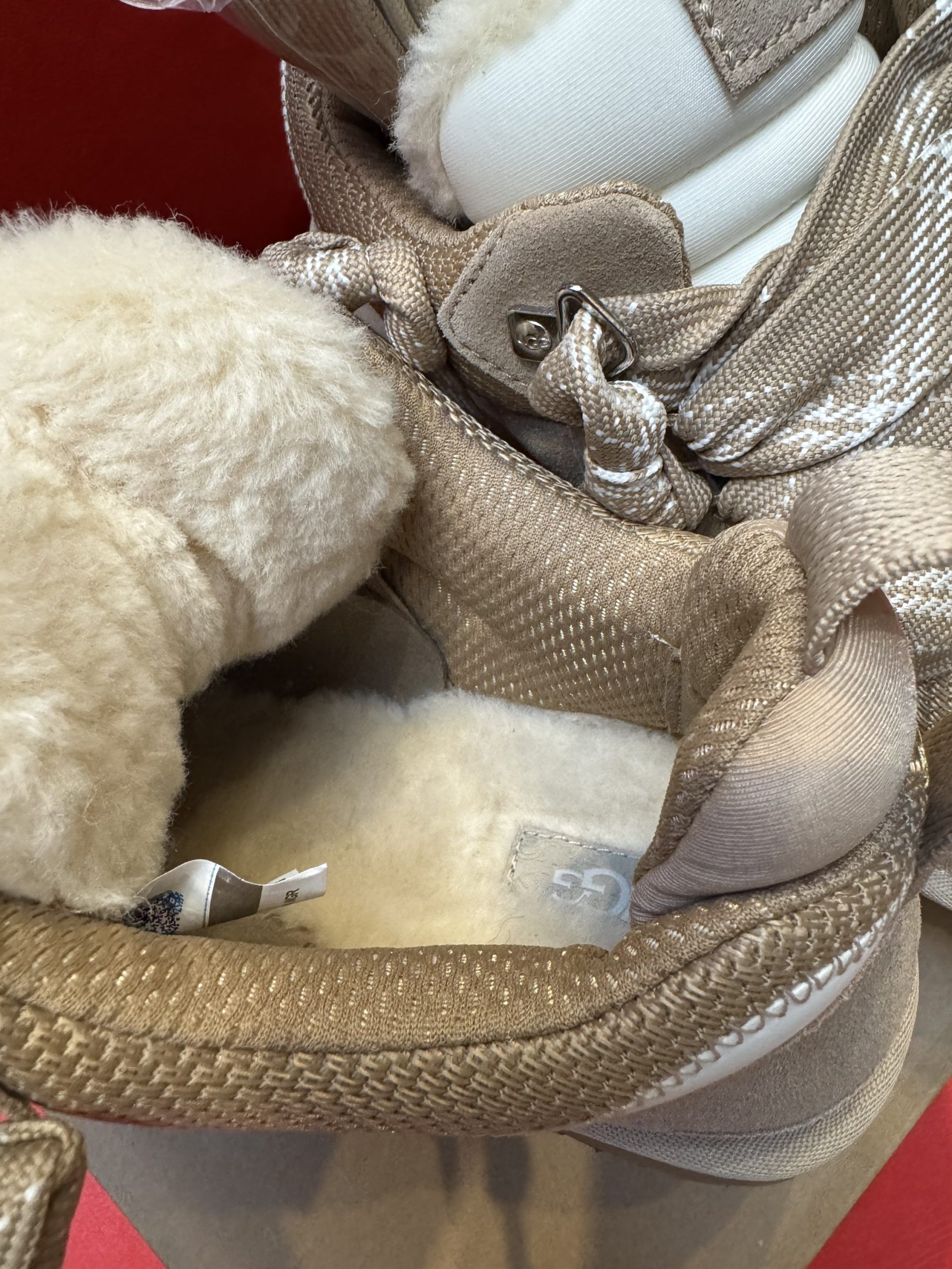 Tan UGG sneaker on a cardboard box with the sole of a matching shoe in the background, resting on a red surface.