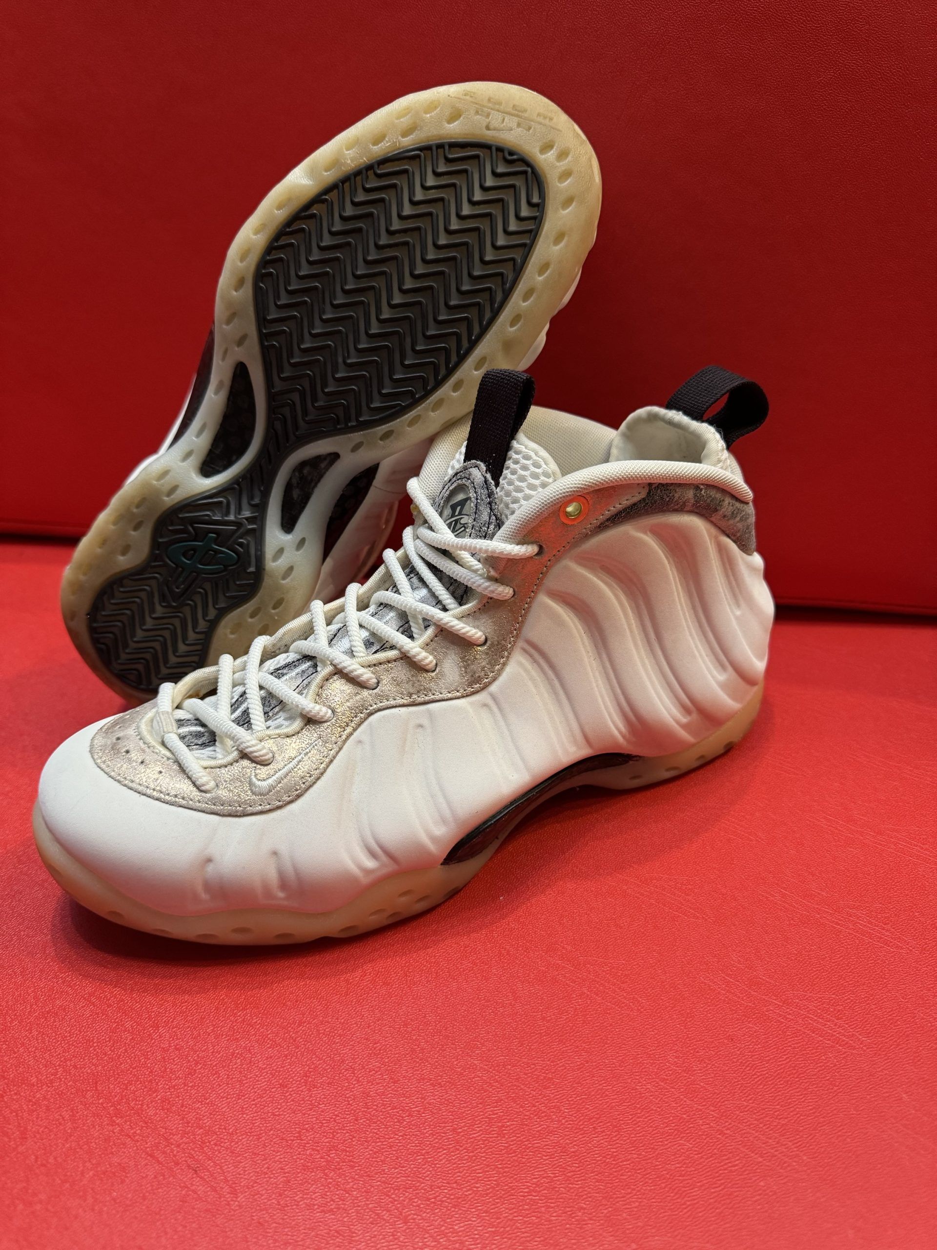 White and gold Nike Foamposite sneakers, one with sole up, on a red surface.