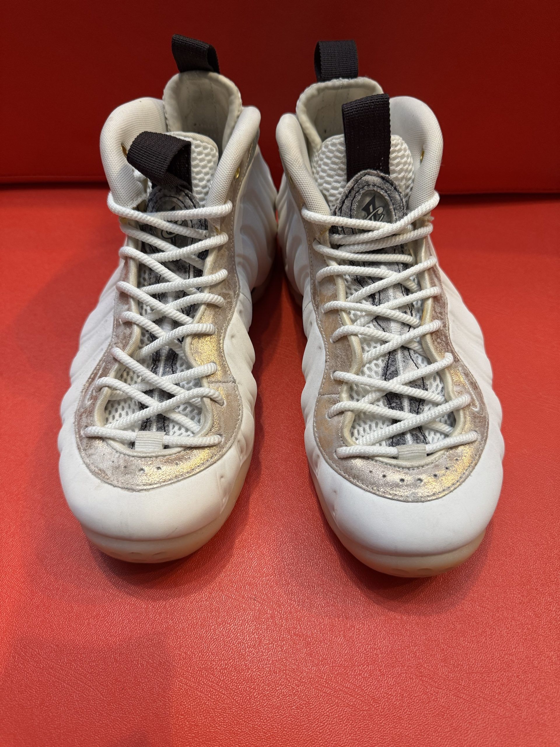 White and gold Nike Foamposite sneakers, one with sole up, on a red surface.