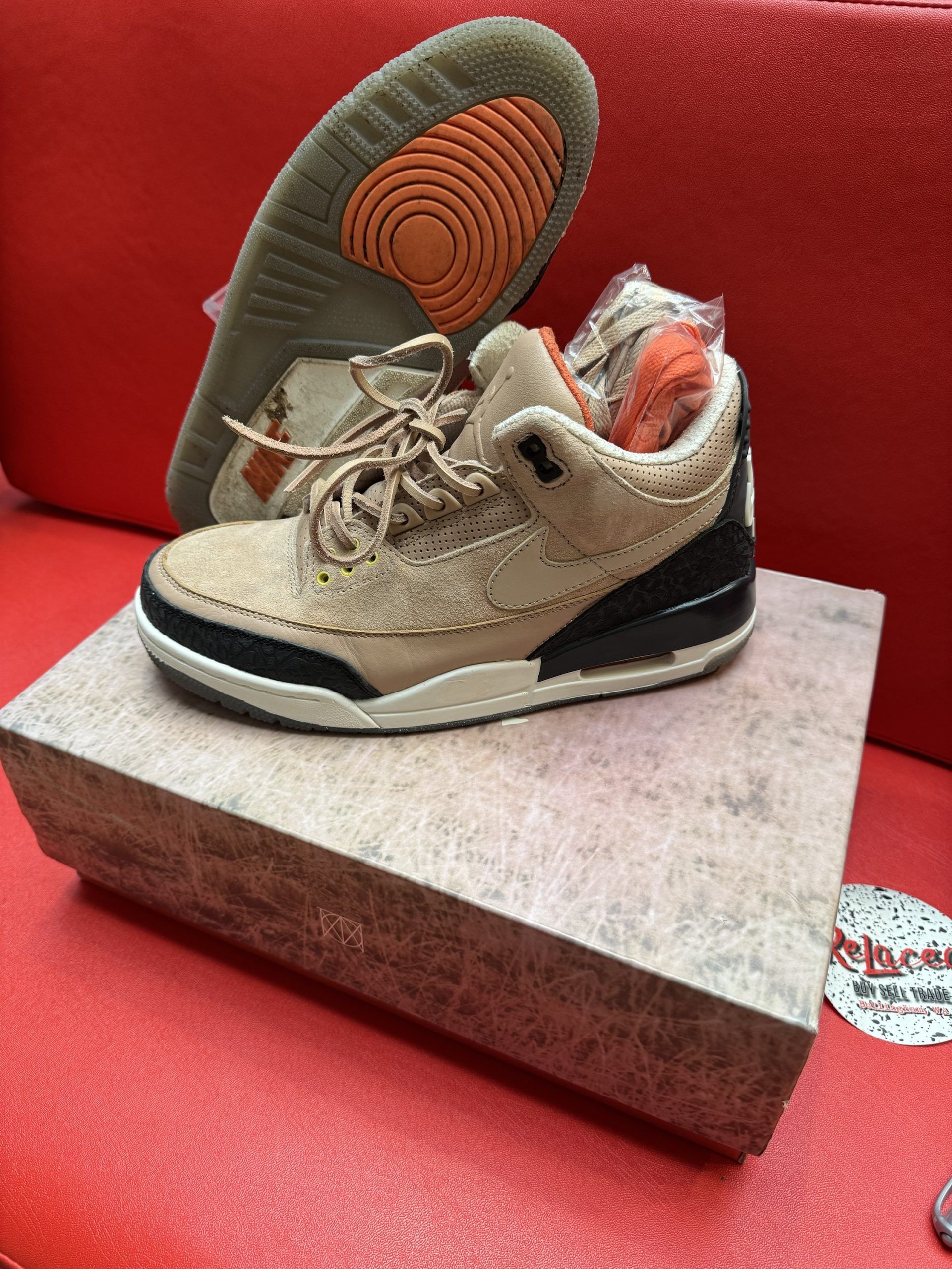 Tan Air Jordan 3 sneakers on a wooden box with a red background. The sole is visible.