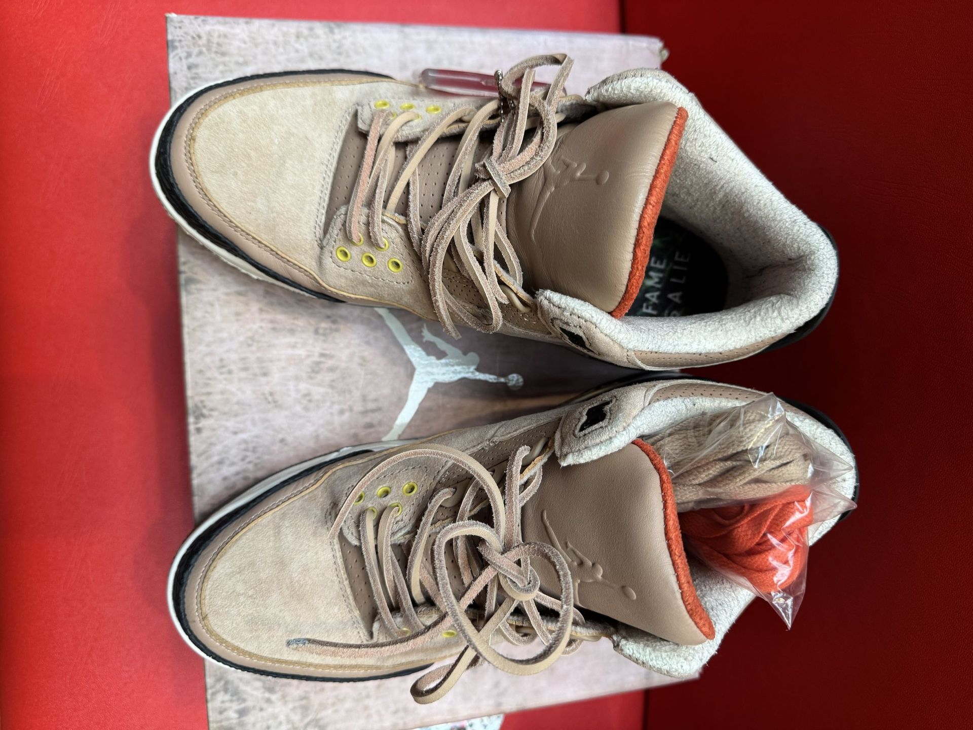 Tan Air Jordan 3 sneakers on a wooden box with a red background. The sole is visible.