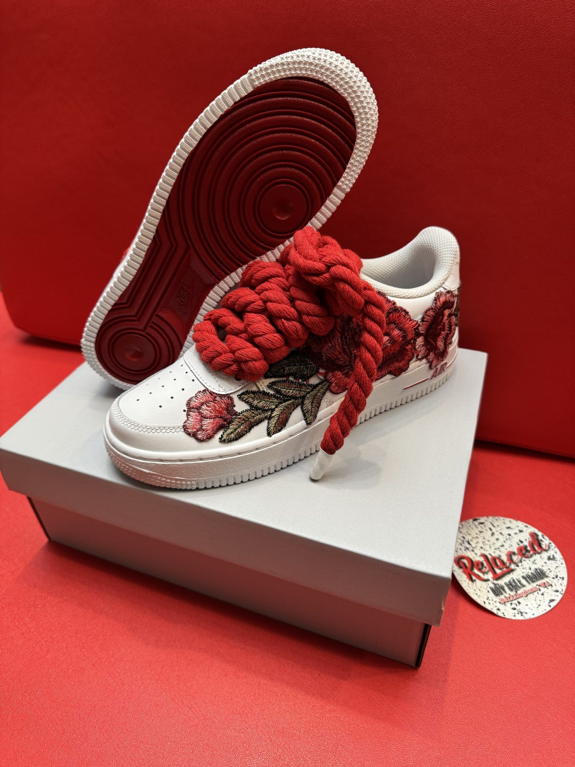 White sneakers with red soles, floral embellishments, and large red laces on a box against a red backdrop.