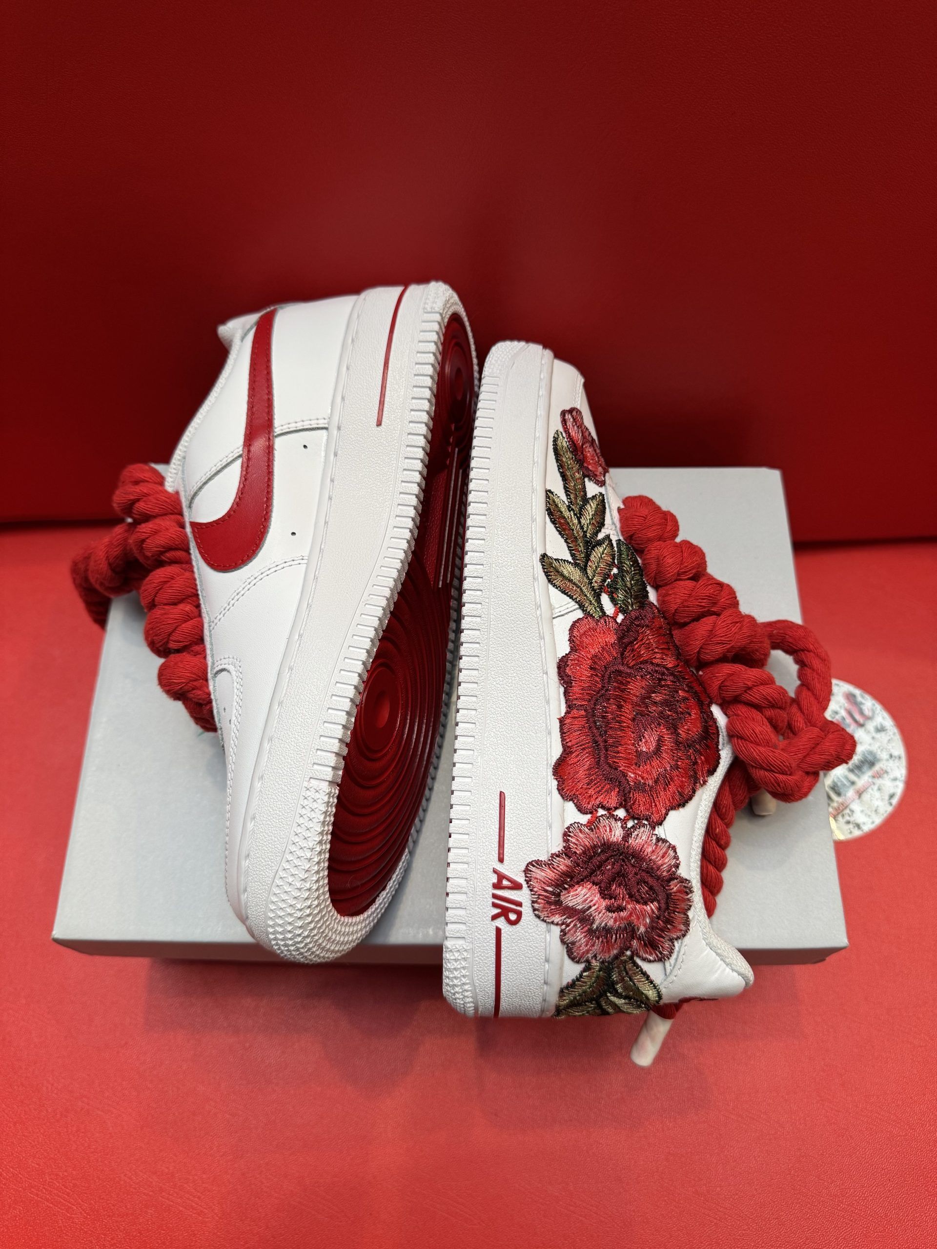 White sneakers with red soles, floral embellishments, and large red laces on a box against a red backdrop.