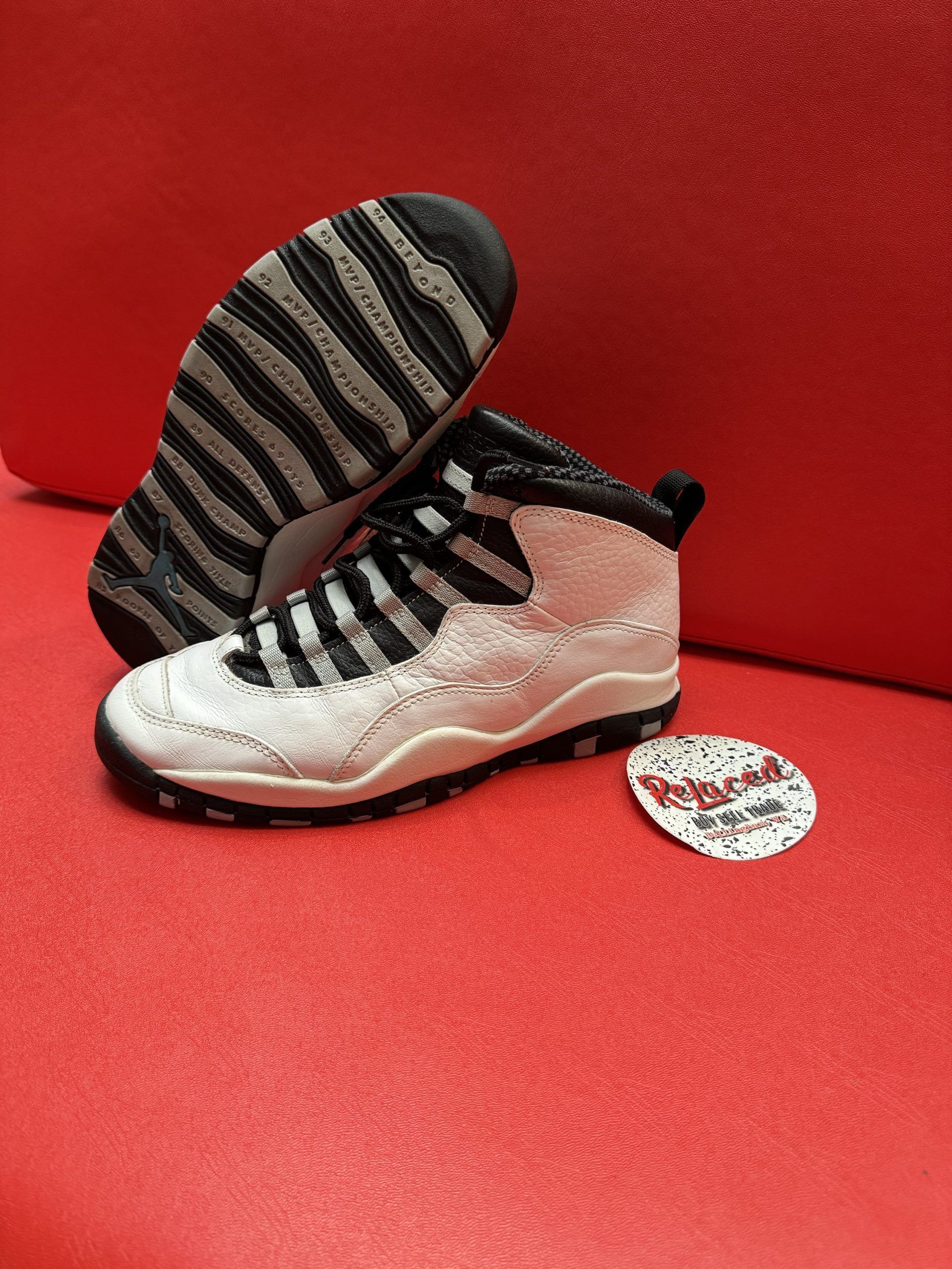 White and black Air Jordan 10 sneakers on a red surface, sole visible, with a round logo.