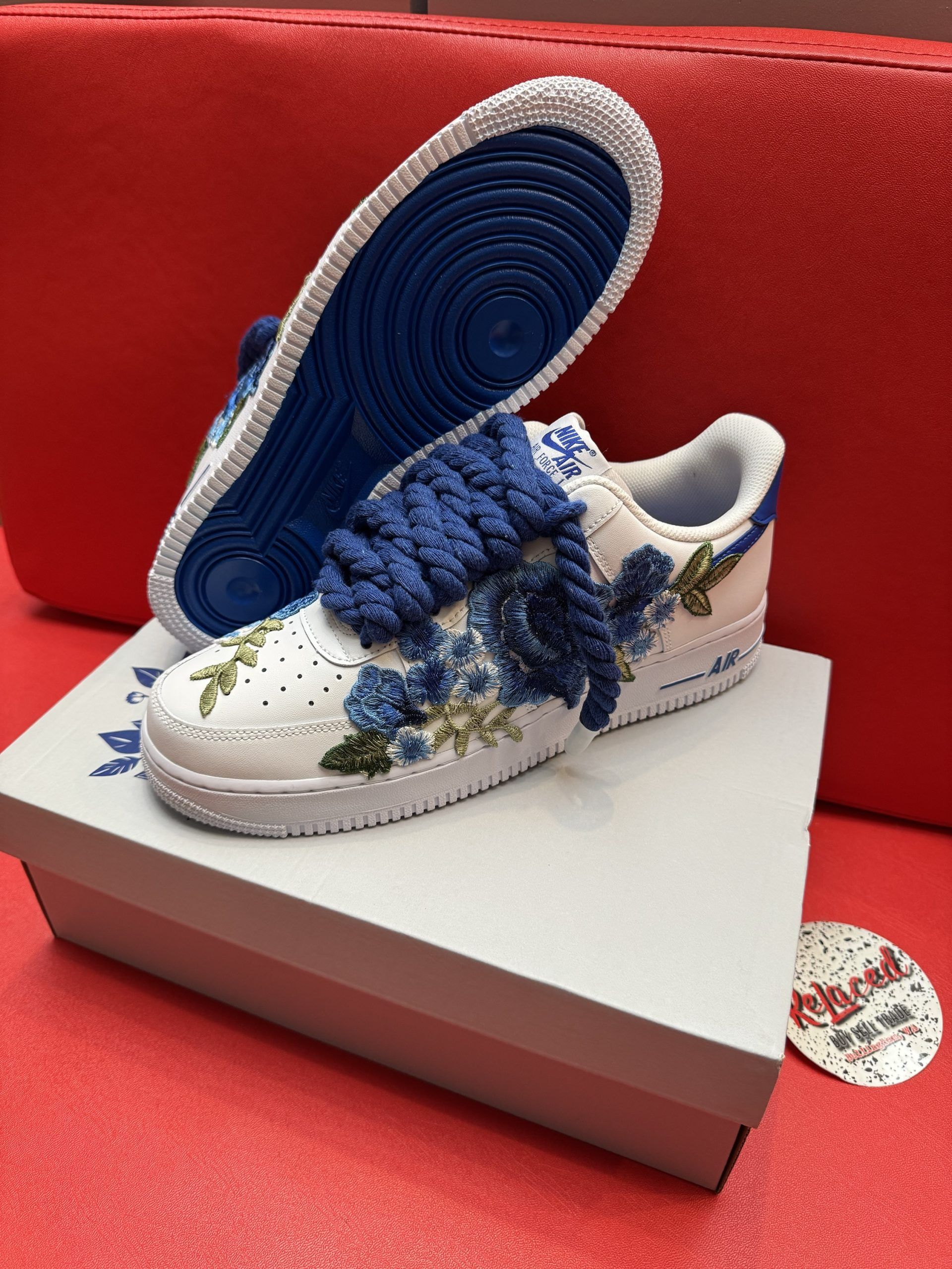 White sneakers with blue floral embroidery and laces, on a white shoebox, red background.