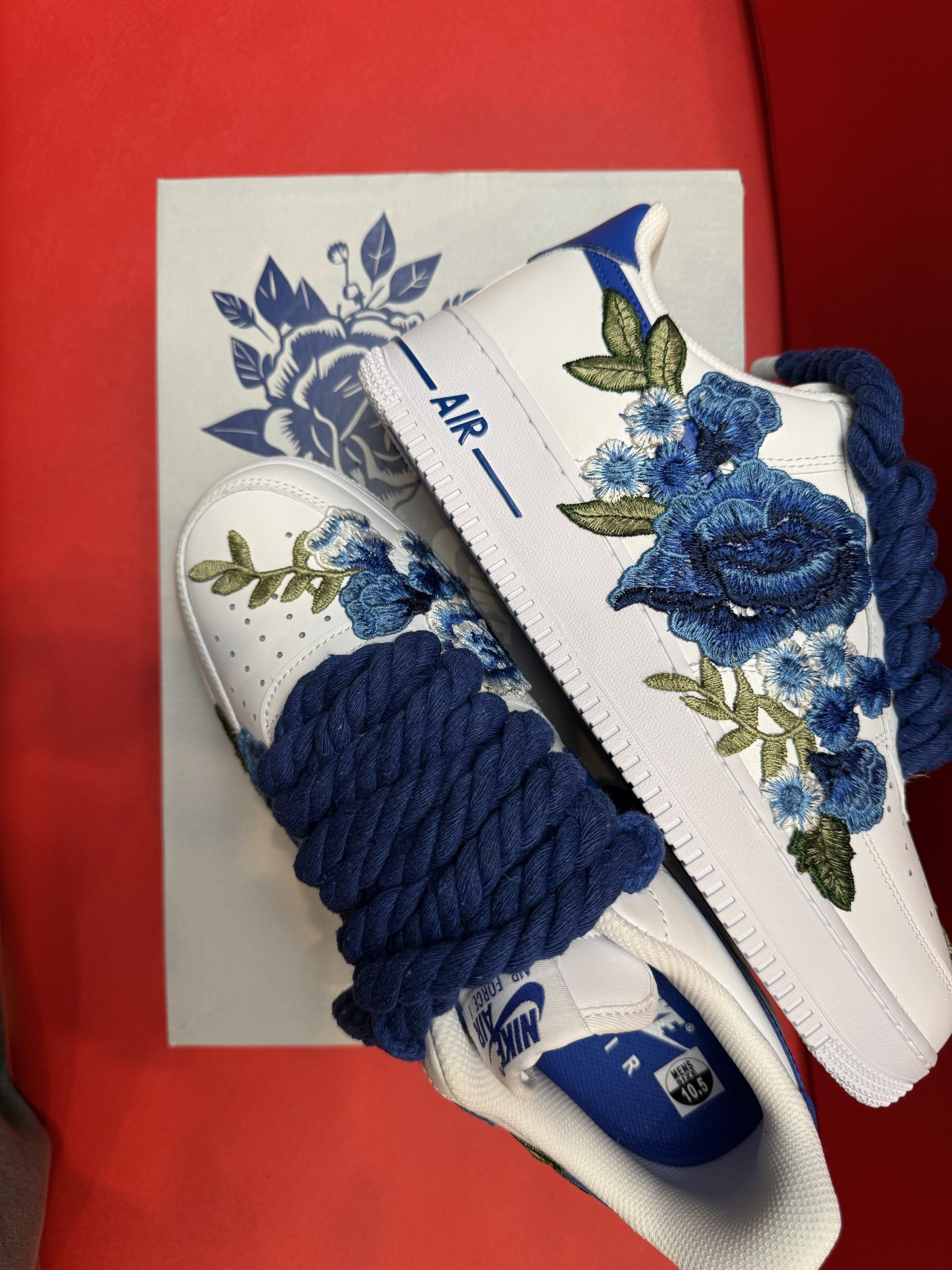 White sneakers with blue floral embroidery and laces, on a white shoebox, red background.