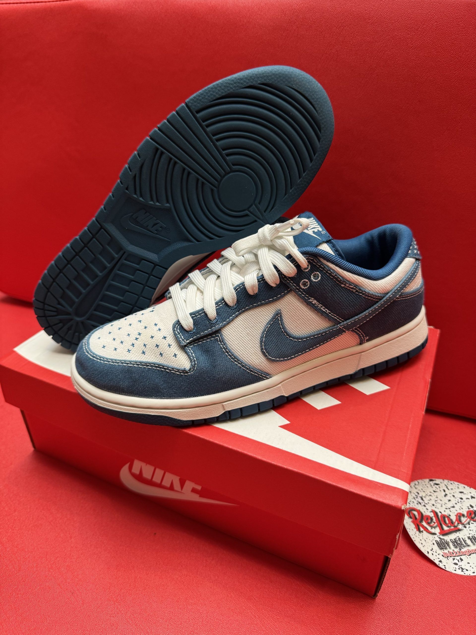 Nike Dunk low-top sneaker in denim, white swoosh, blue sole, on a red box, red backdrop.