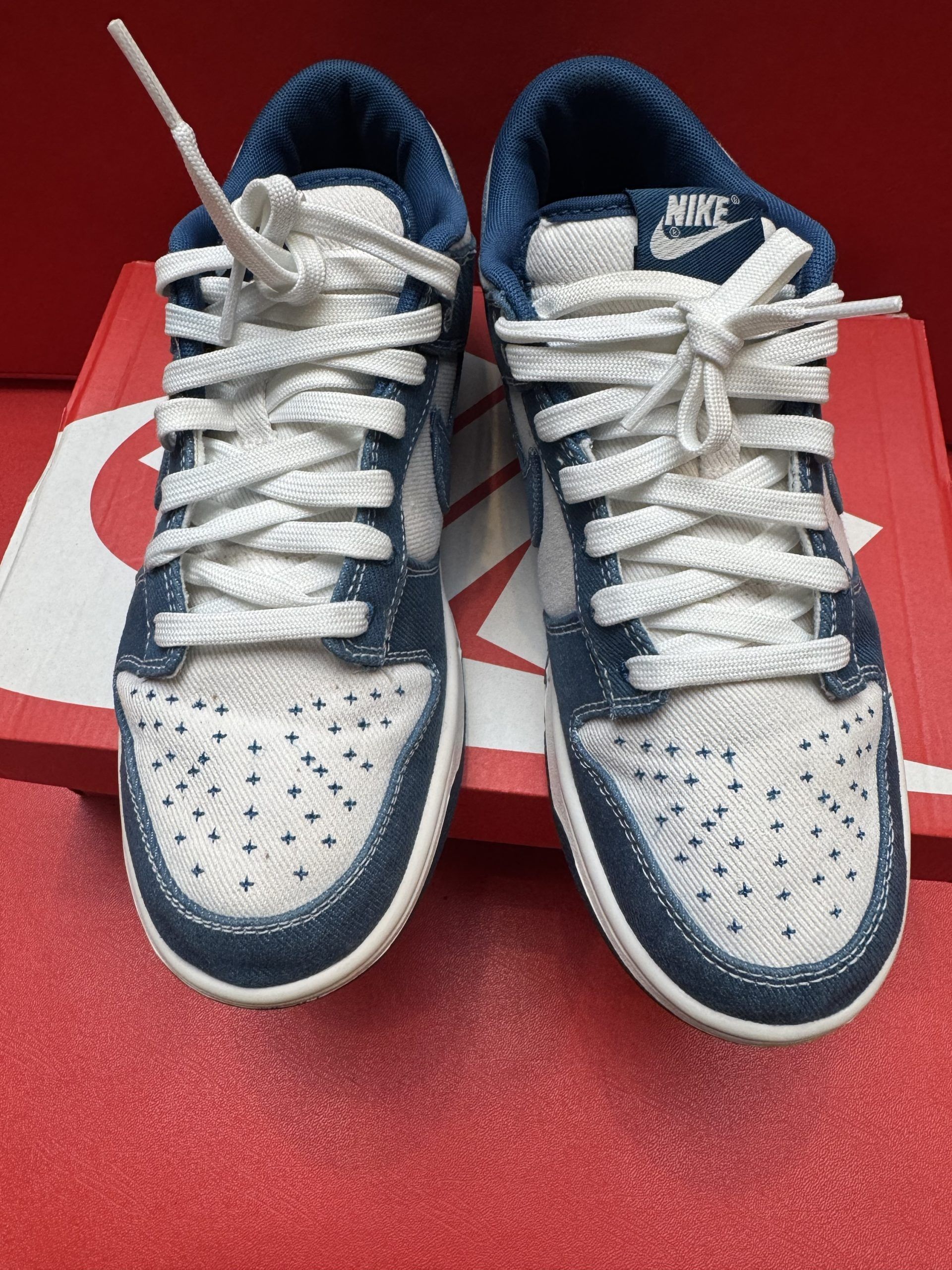 Nike Dunk low-top sneaker in denim, white swoosh, blue sole, on a red box, red backdrop.